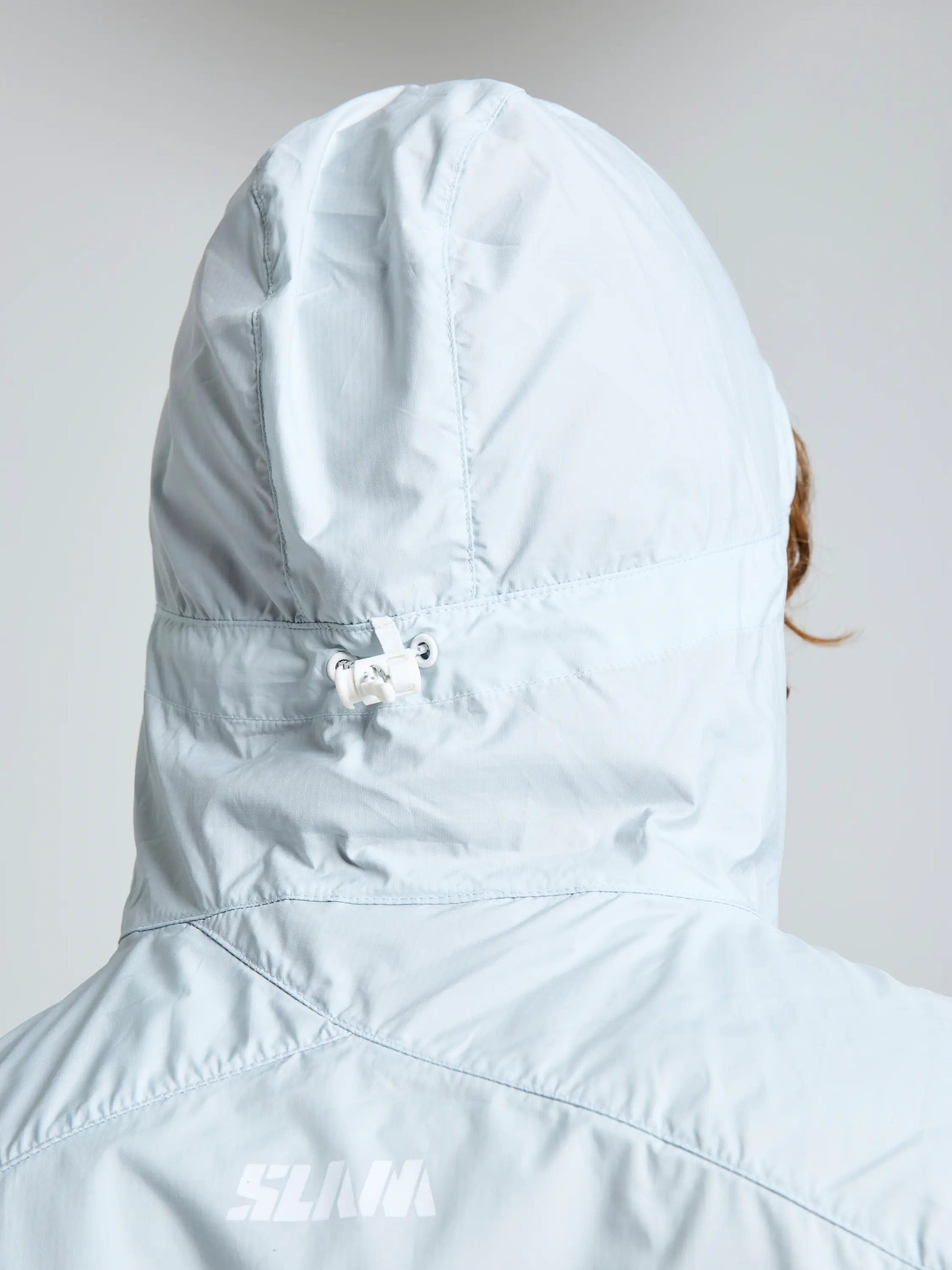 A person is shown from the back wearing a light gray ACT PACKLITE JACKET by SLAM. The windproof hood covers their head, revealing a bit of brown hair on the right side. "SLAM" is printed on this men's jacket.