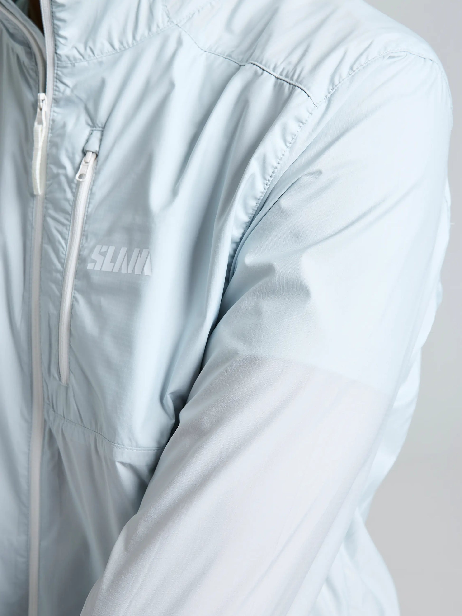 A close-up of a person wearing the SLAM ACT PACKLITE JACKET, a light blue lightweight jacket with a white zipper, small chest pocket, and partially visible white logo text on the left chest.