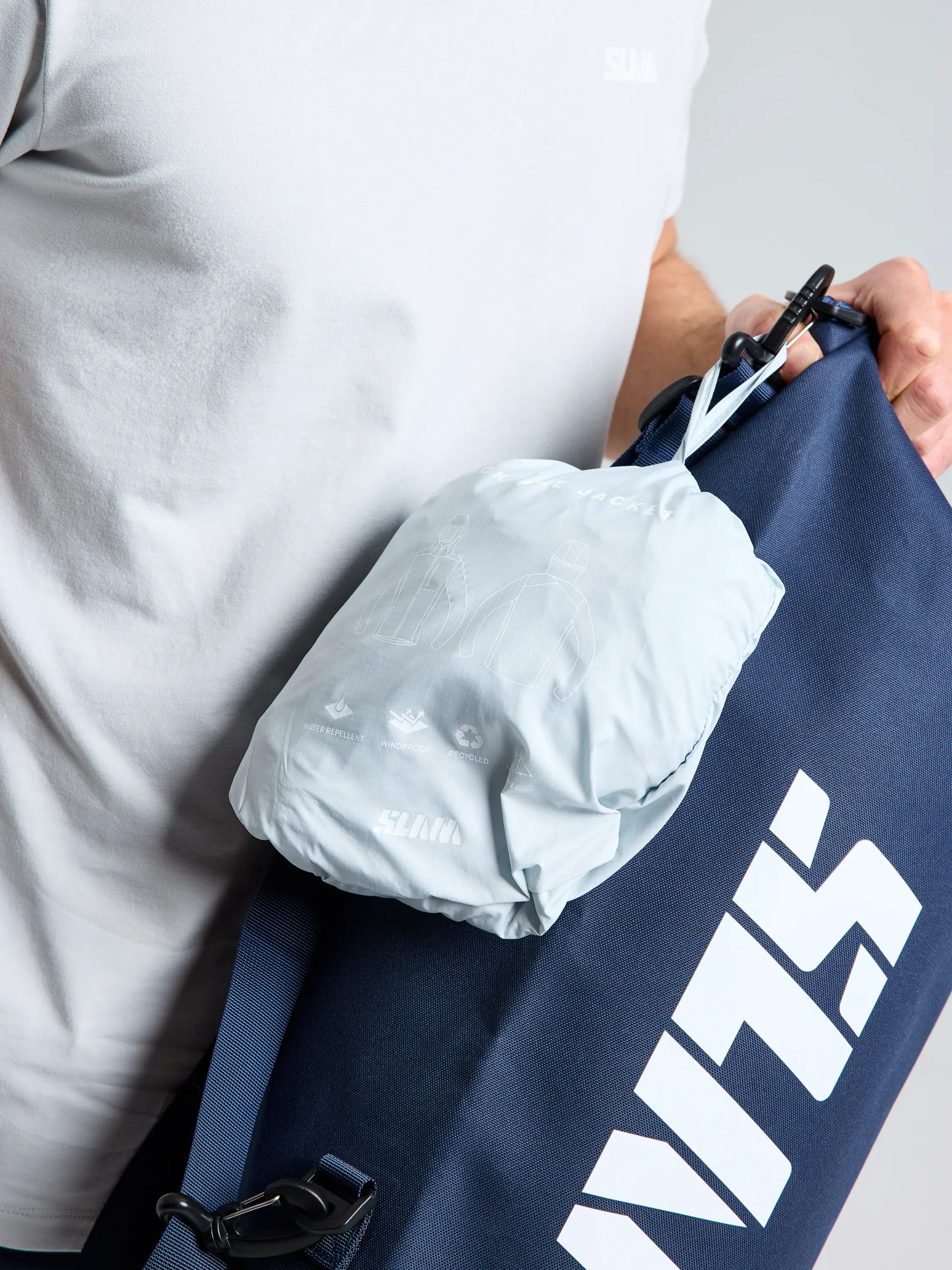A person wears a light grey shirt and holds a navy blue SLAM bag with bold white "SLAM" letters, plus an attached light grey pouch—ideal for carrying the SLAM ACT PACKLITE JACKET on the go.
