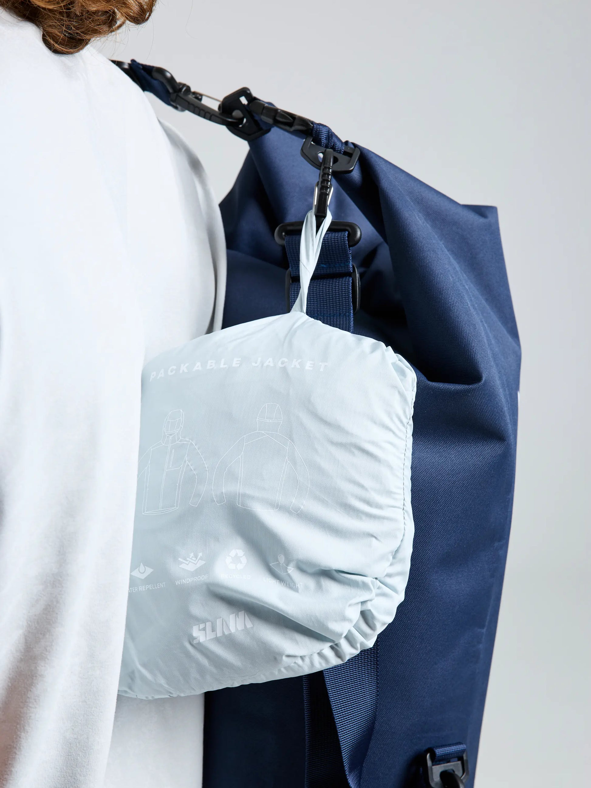 A person in a white shirt carries a dark blue bag over their shoulder. Attached is the SLAM ACT PACKLITE JACKET—a light gray, compact jacket with printed graphics and text on its surface.