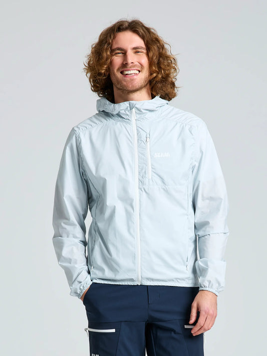 A curly-haired person smiles warmly in the SLAM ACT PACKLITE JACKET—a light blue, front-zip, packable jacket—paired with navy pants. The lightweight piece stands out boldly against a plain light background.