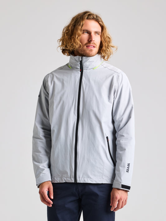 A man with curly hair is wearing the SLAM CREW WARM HOODED JACKET in Glacier Grey, perfect for winter sailing, featuring a high collar and black zipper accents. The waterproof jacket displays "WITTE" on the sleeve. He pairs it with dark pants while looking slightly to the side.