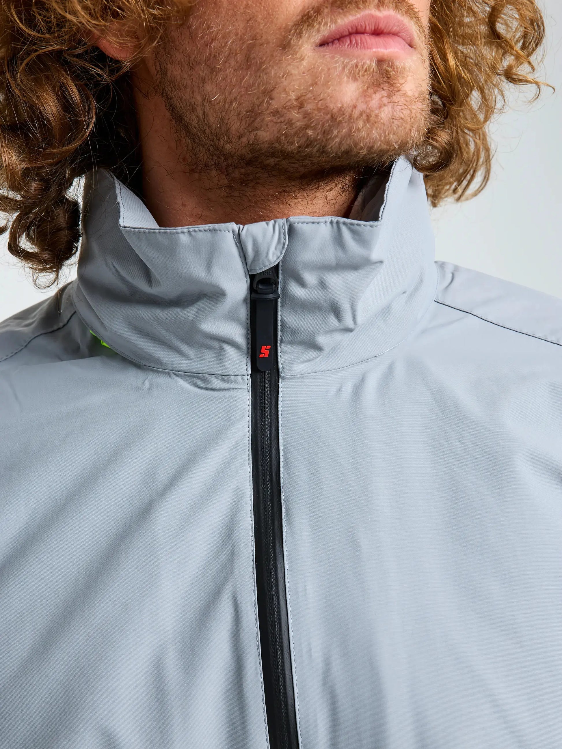 A close-up of a person with curly hair wearing the SLAM CREW WARM HOODED JACKET in light gray, featuring a high collar and black zipper. The face is partially visible, drawing attention to the jacket’s neckline and practical design—perfect for winter sailing.