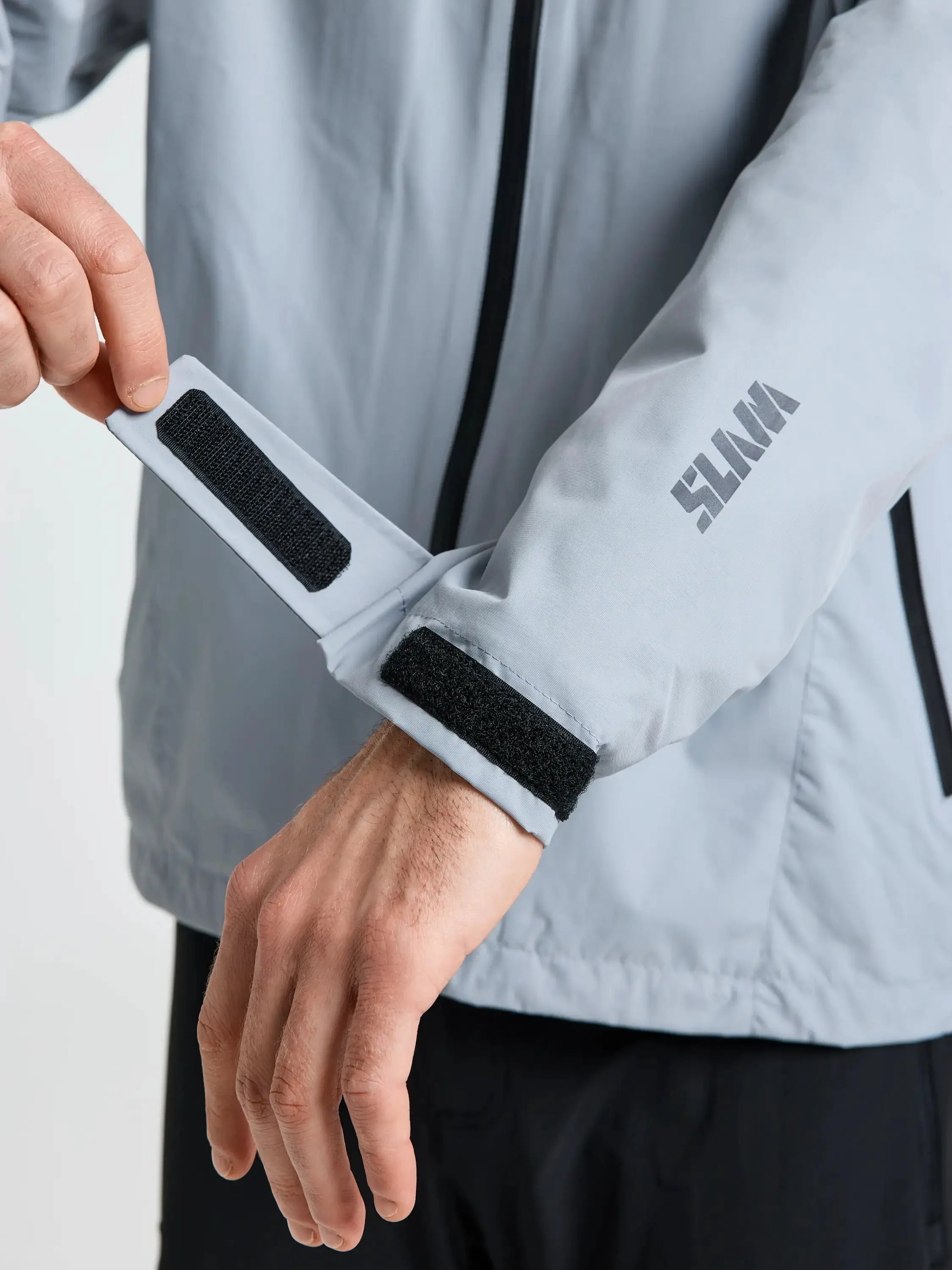 A person adjusts the Velcro cuff strap on the sleeve of a light gray CREW WARM HOODED JACKET by SLAM, ideal as a winter sailing jacket. The brand logo is printed on the forearm, and only the torso and arms are visible.