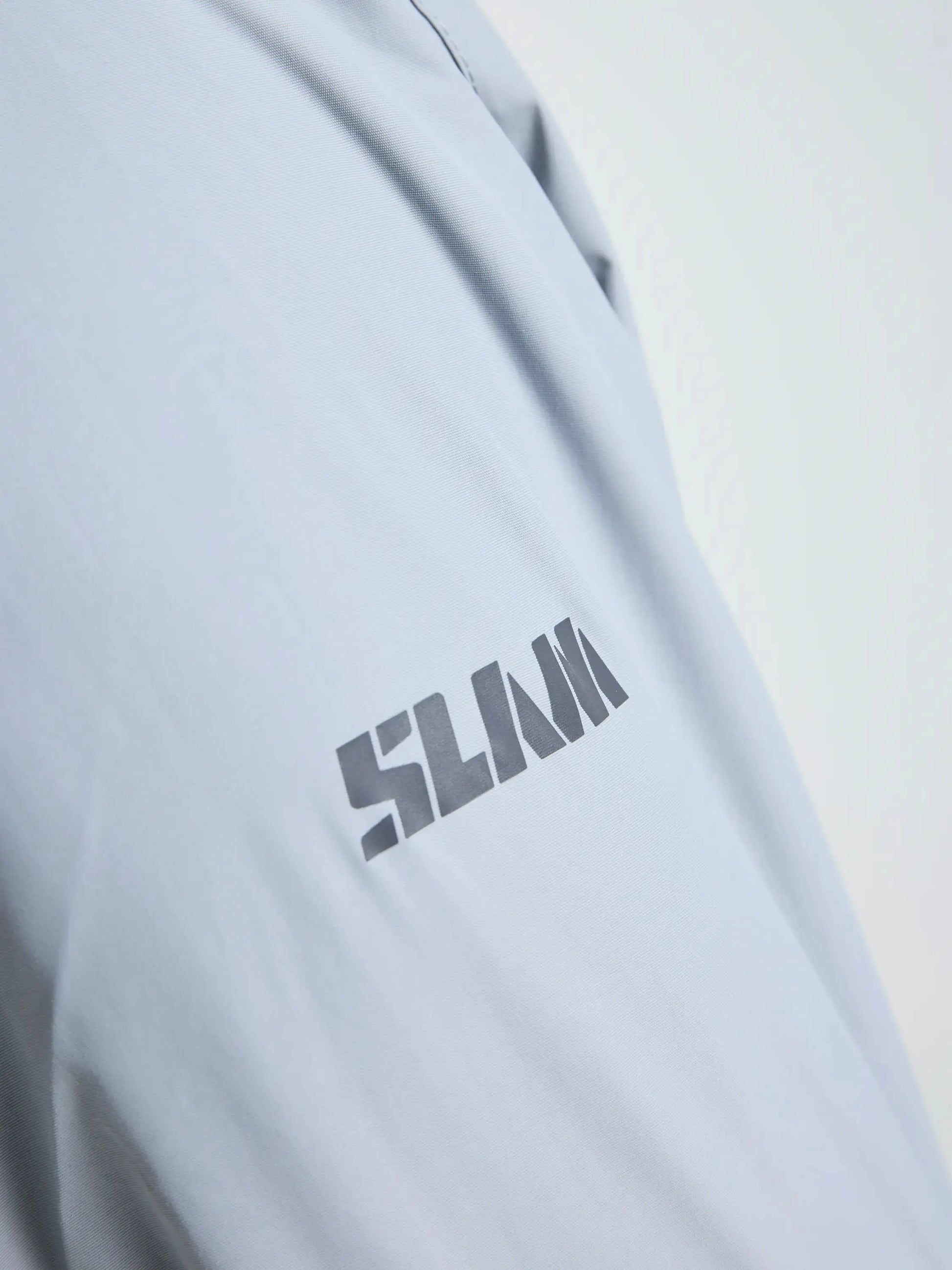 Close-up of a light gray fabric—part of the SLAM CREW WARM HOODED JACKET—featuring the brand name “SLAM” in bold, dark, block-style letters. Set against a white background, the image emphasizes both the printed text and the texture of this waterproof winter sailing jacket’s material.