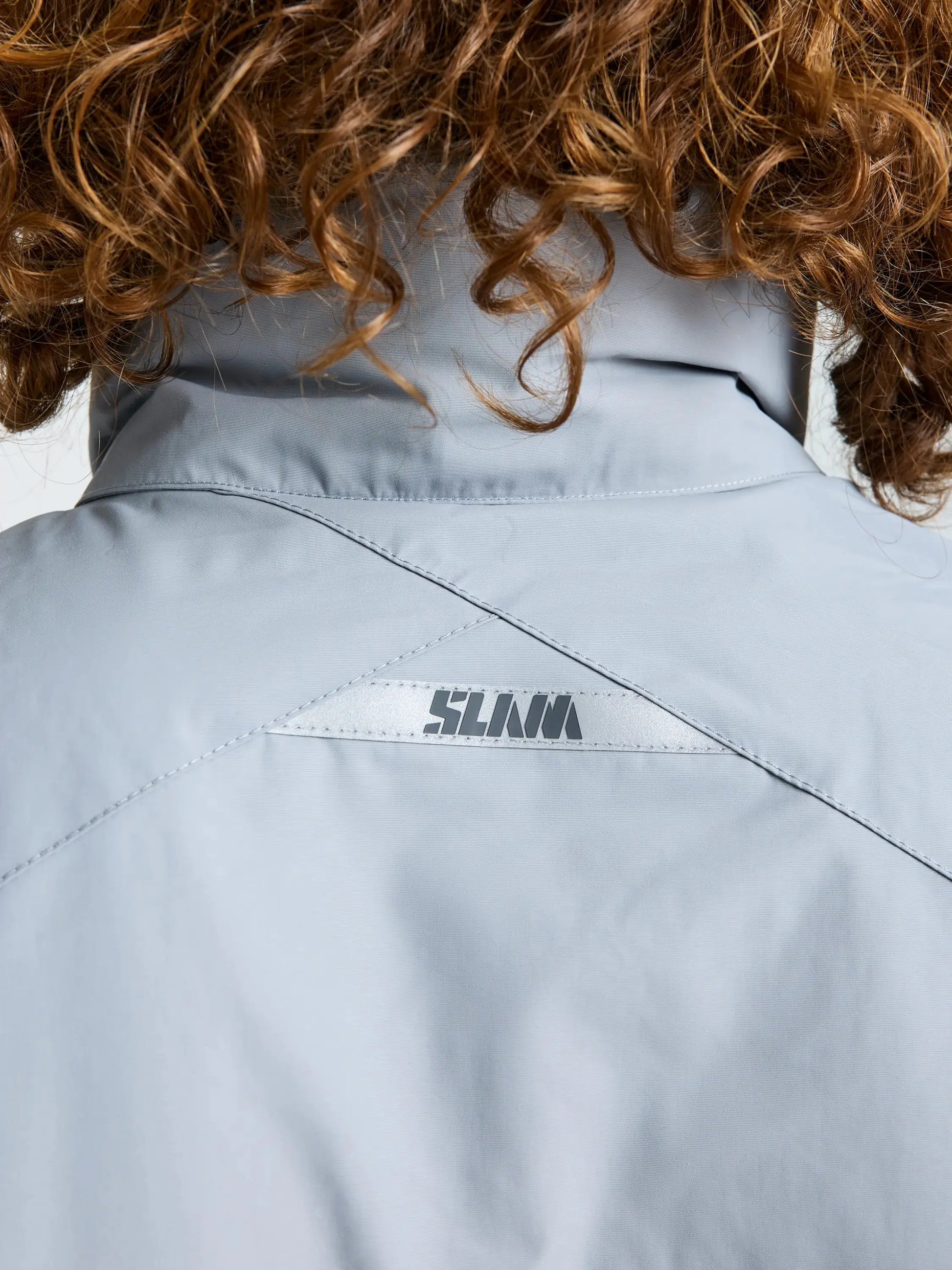 Close-up of the back of a light gray SLAM CREW WARM HOODED JACKET with a small “SLNM” logo printed below the collar. The wearer has curly brown hair.