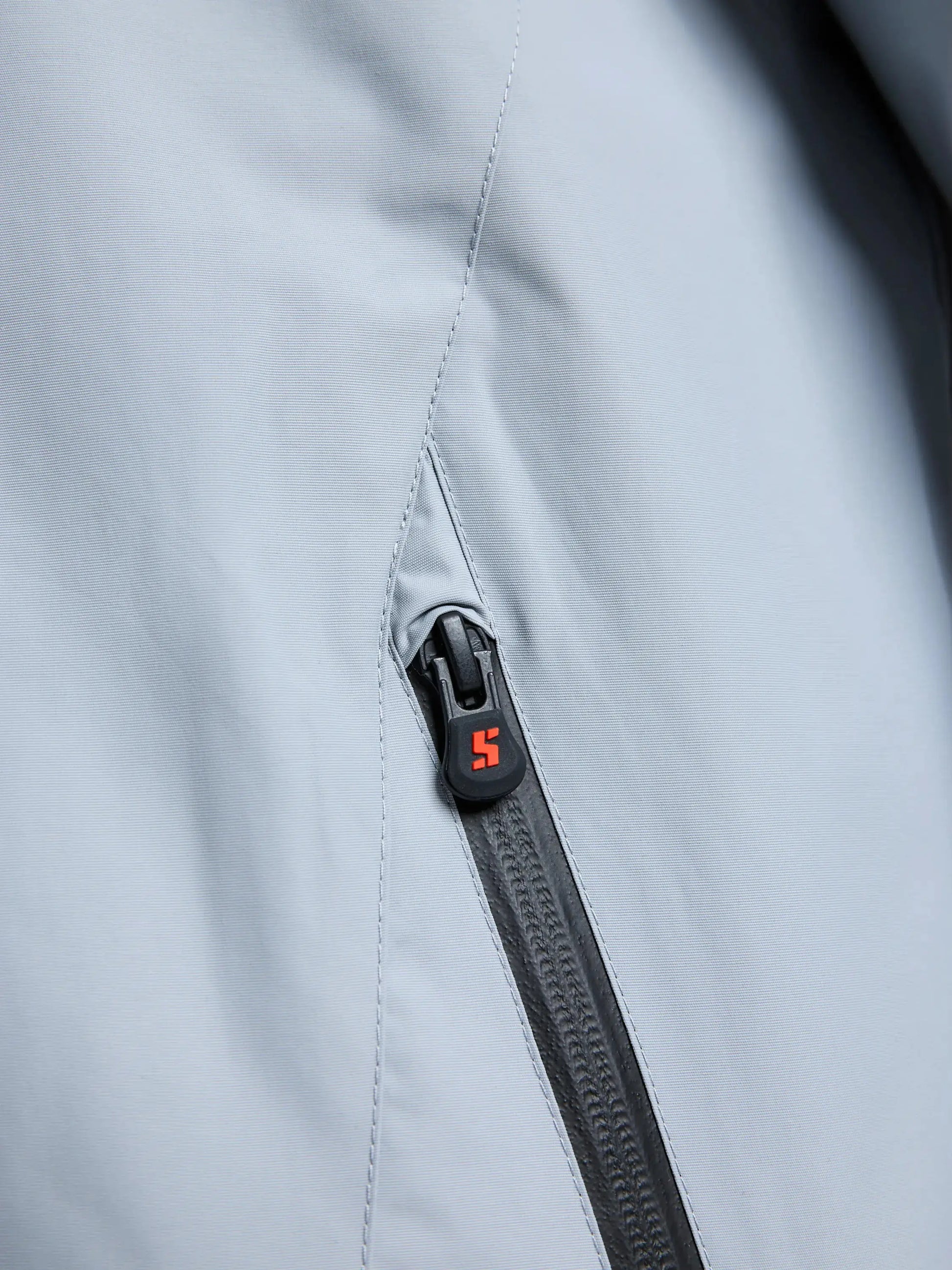Close-up of the SLAM CREW WARM HOODED JACKET in gray, featuring a black zipper with a small red and white “S” logo on the zipper pull. The fabric is smooth, slightly reflective, and has the look of high-performance waterproof material designed for winter sailing.