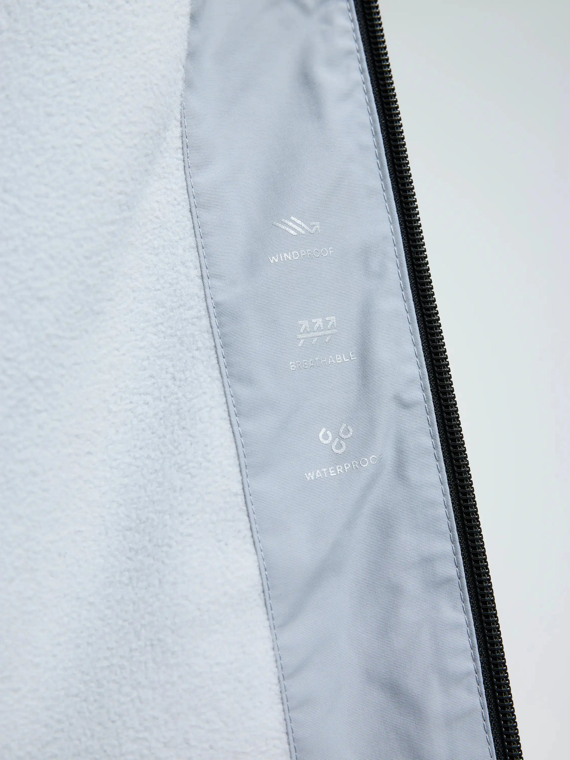 Close-up of the interior of the CREW WARM HOODED JACKET by SLAM in light gray, with icons and text highlighting its windproof, breathable, and waterproof features. The edge of a black zipper appears on the right, emphasizing technical design elements that make this SLAM jacket ideal for harsh winter sailing conditions.