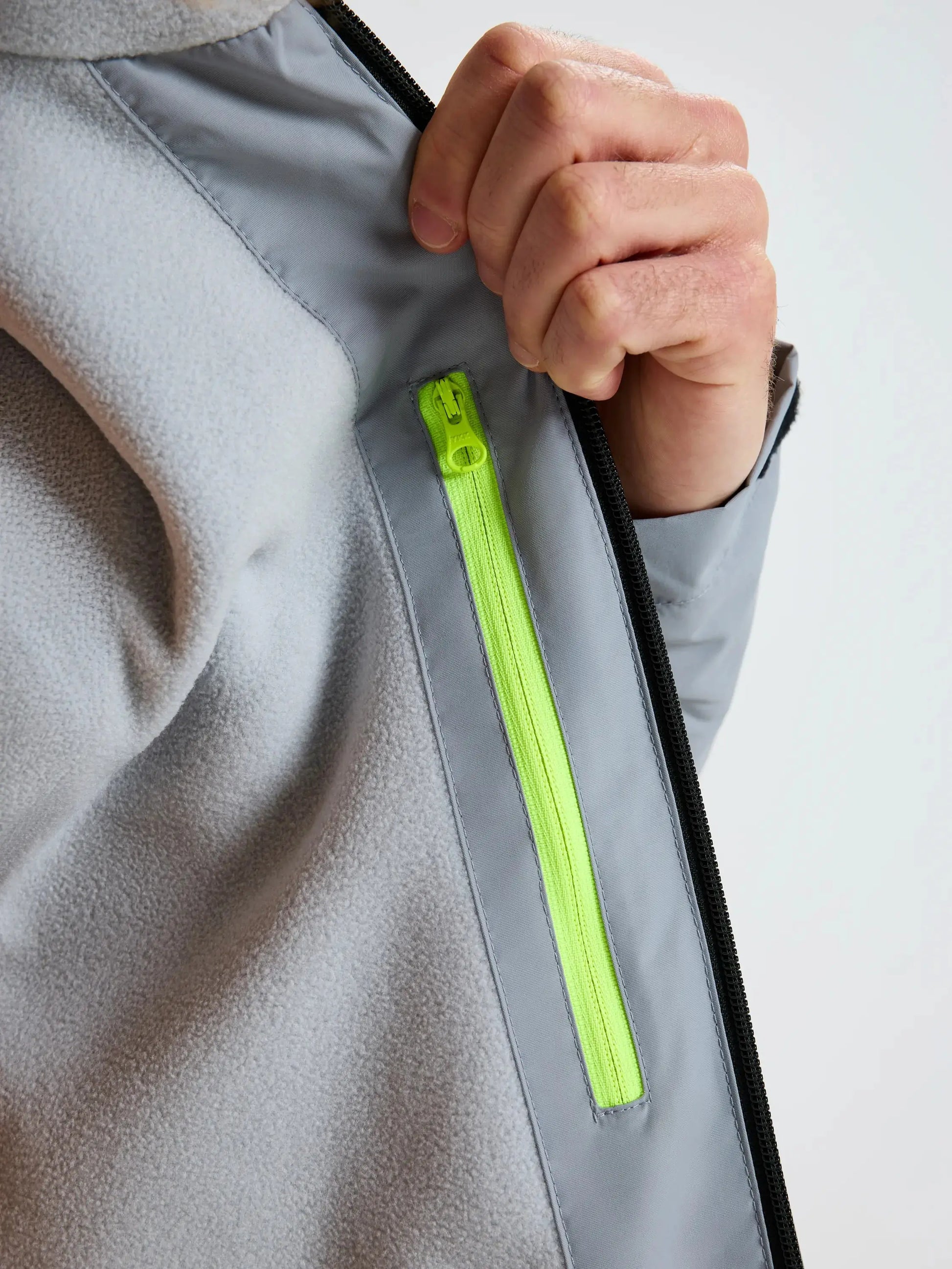 A person pulls open the SLAM CREW WARM HOODED JACKET in light gray, revealing an inner chest pocket with a vertical neon yellow zipper. The jacket appears to be made of a soft, fleece-like material.