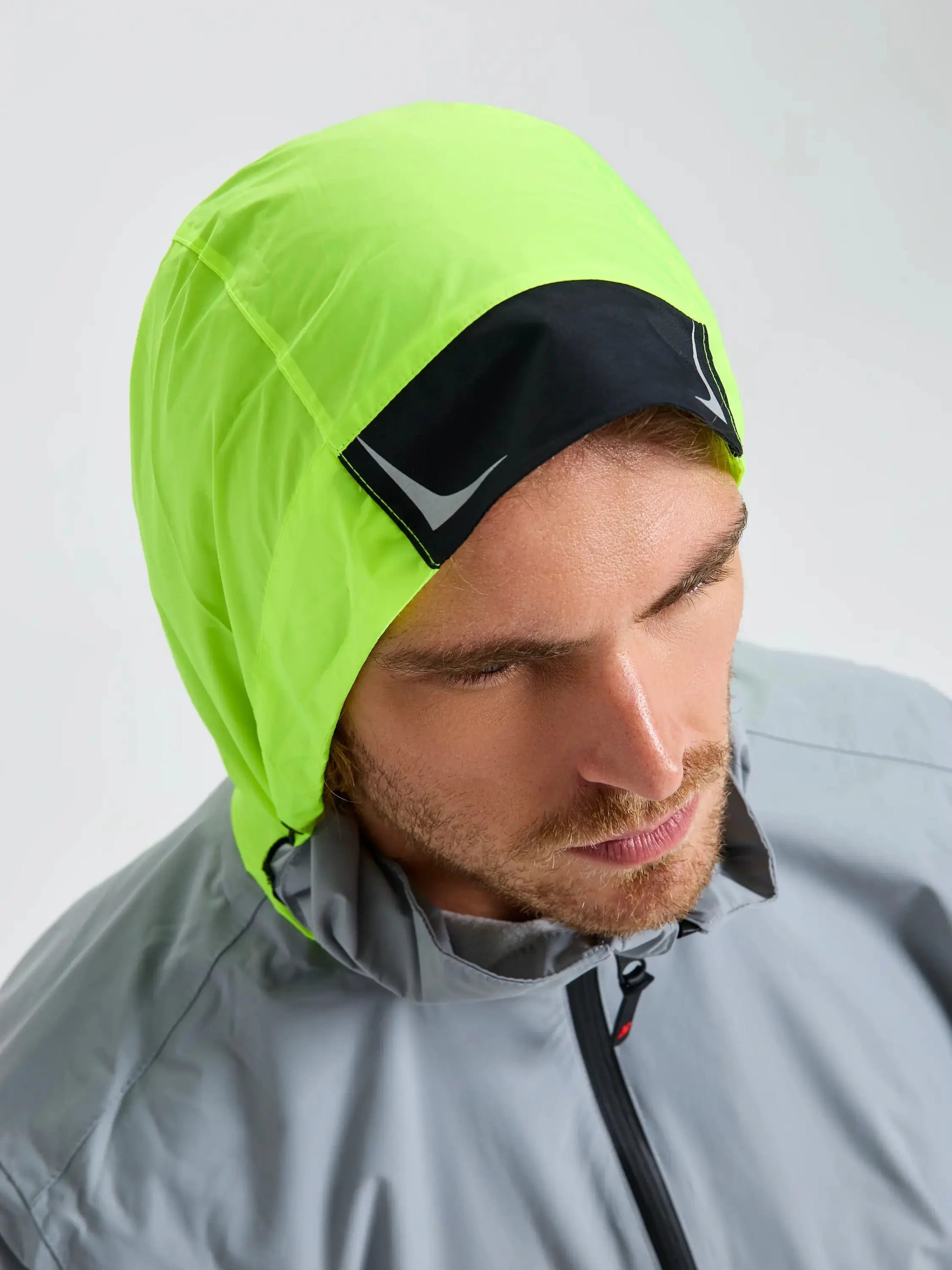 A man wearing the SLAM CREW WARM HOODED JACKET in light gray, featuring a neon yellow hood and a black front patch, looks downward against a plain, light background.