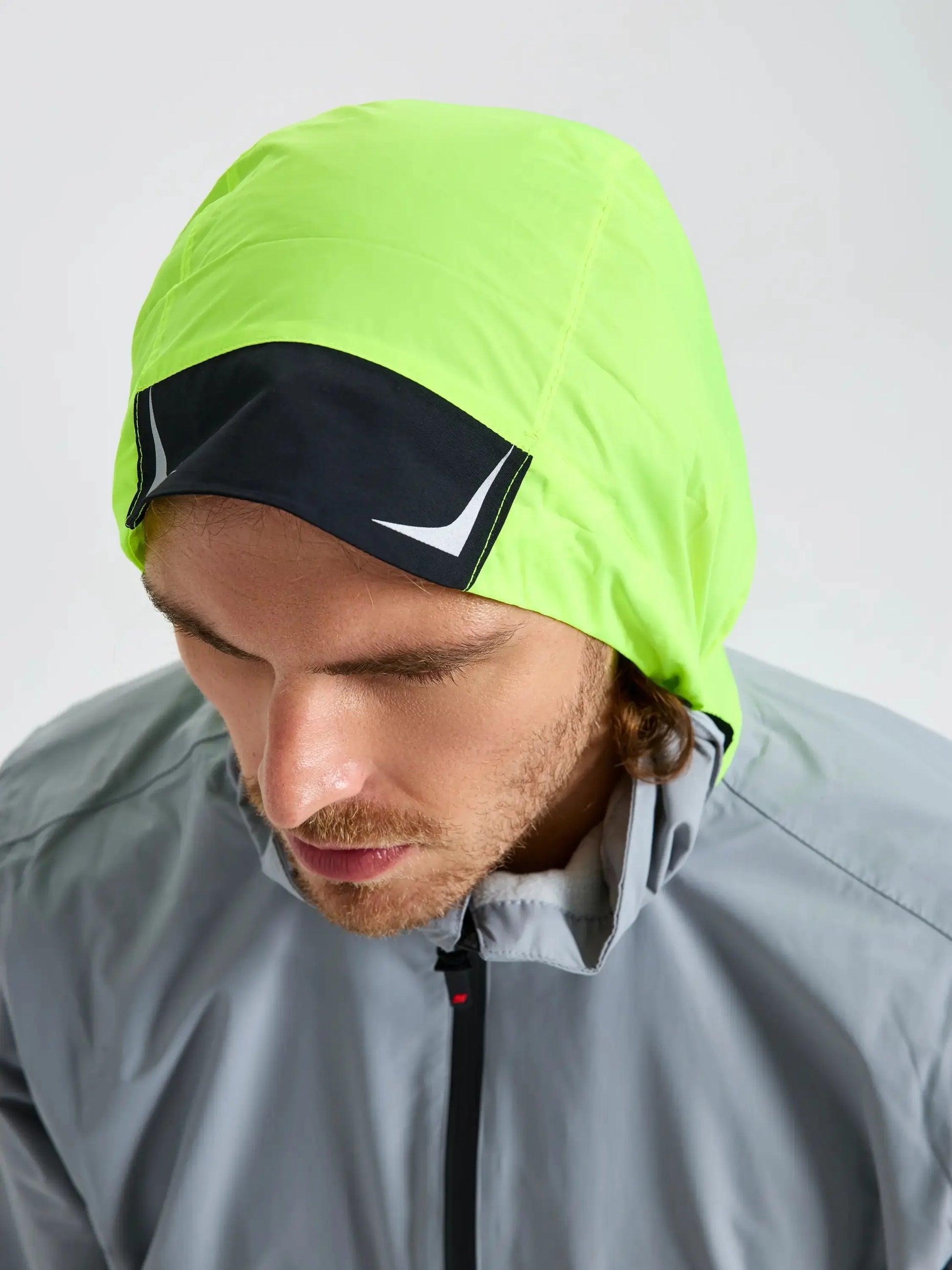 A person wearing the SLAM CREW WARM HOODED JACKET in gray, which features a lime green hood and a black and white patch on the front, is looking downward against a plain background.