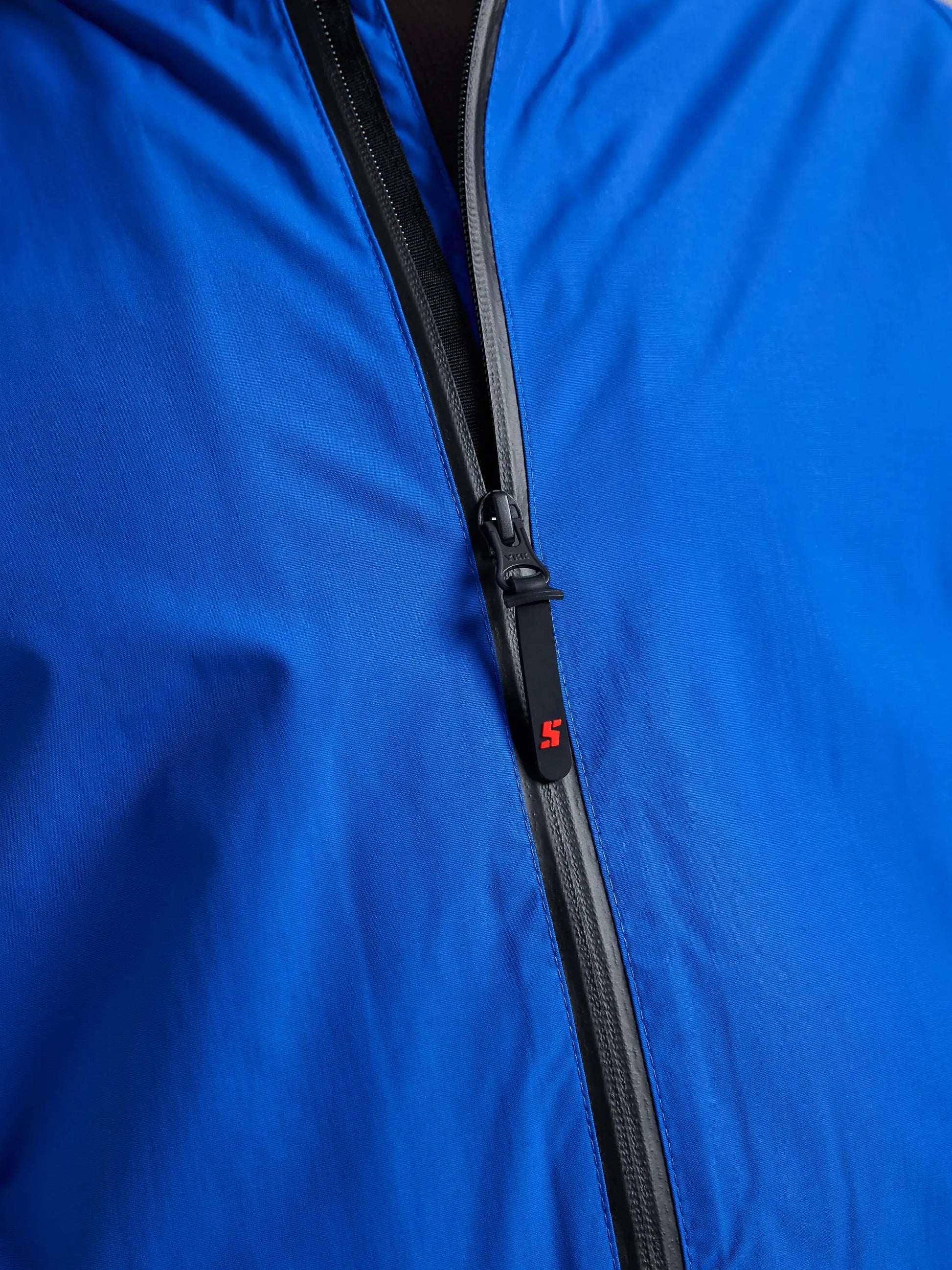 Close-up of the SLAM CREW WARM HOODED JACKET in blue, featuring a black zipper that is partially unzipped. The zipper pull tab displays a small red “S” logo. The jacket’s smooth fabric is visible, along with a glimpse of the black lining near the top.