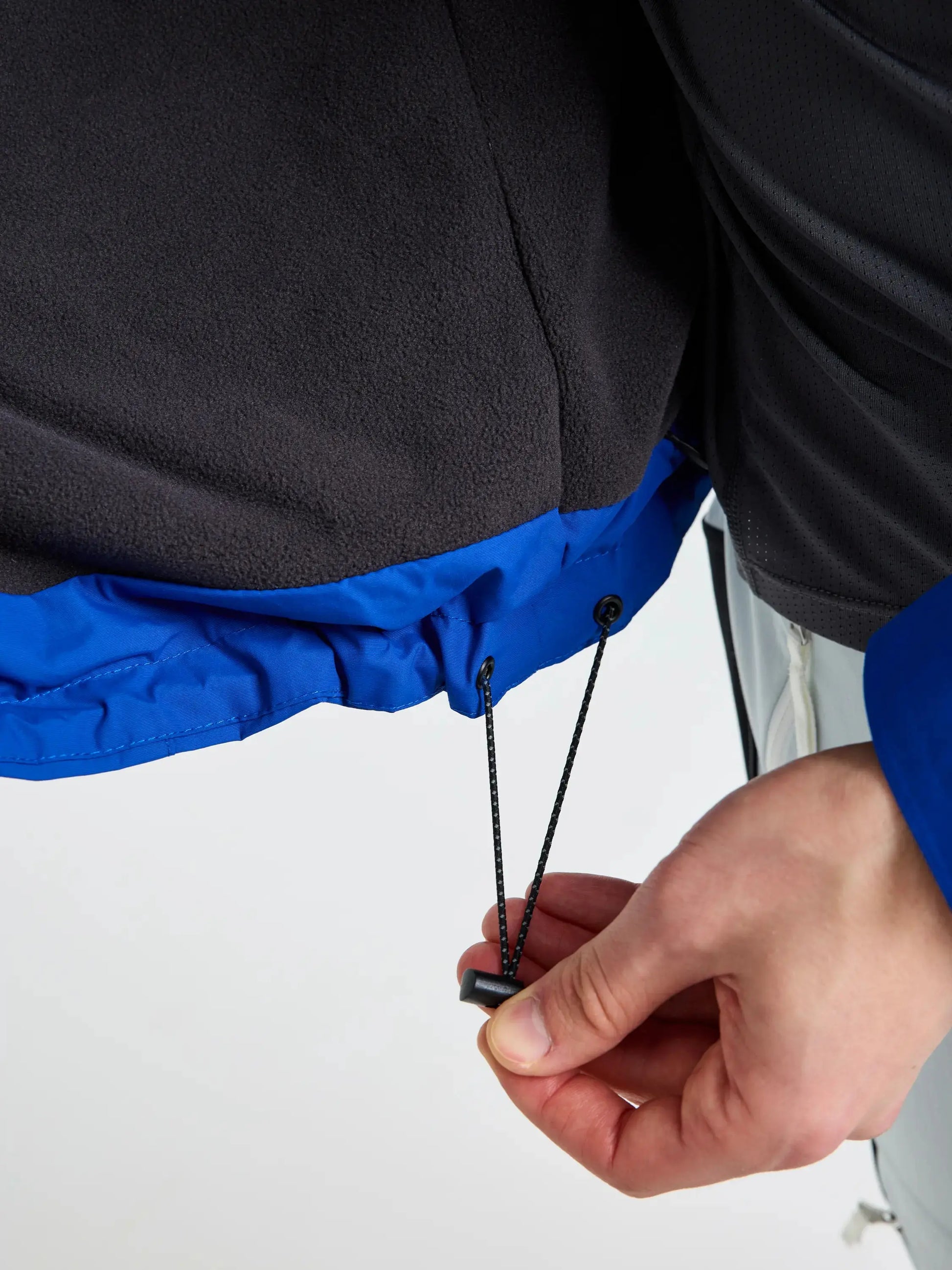 A close-up of a hand adjusting the elastic drawstring hem of the SLAM CREW WARM HOODED JACKET in blue and black, tightening it with a black plastic cord lock. The background is plain and light-colored.
