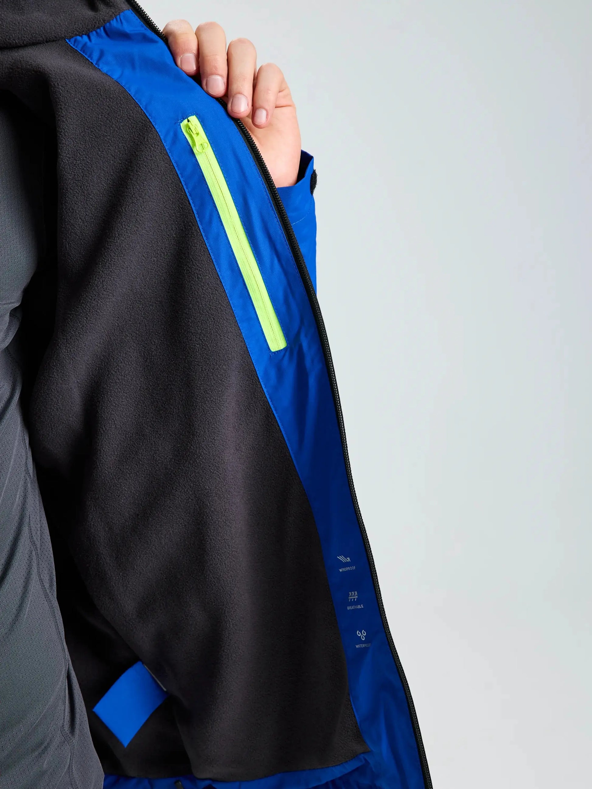 A person holds open the SLAM CREW WARM HOODED JACKET in blue and black, revealing a neon yellow zipper on an inside pocket. This jacket features technical outdoor fabric designed for waterproof protection.