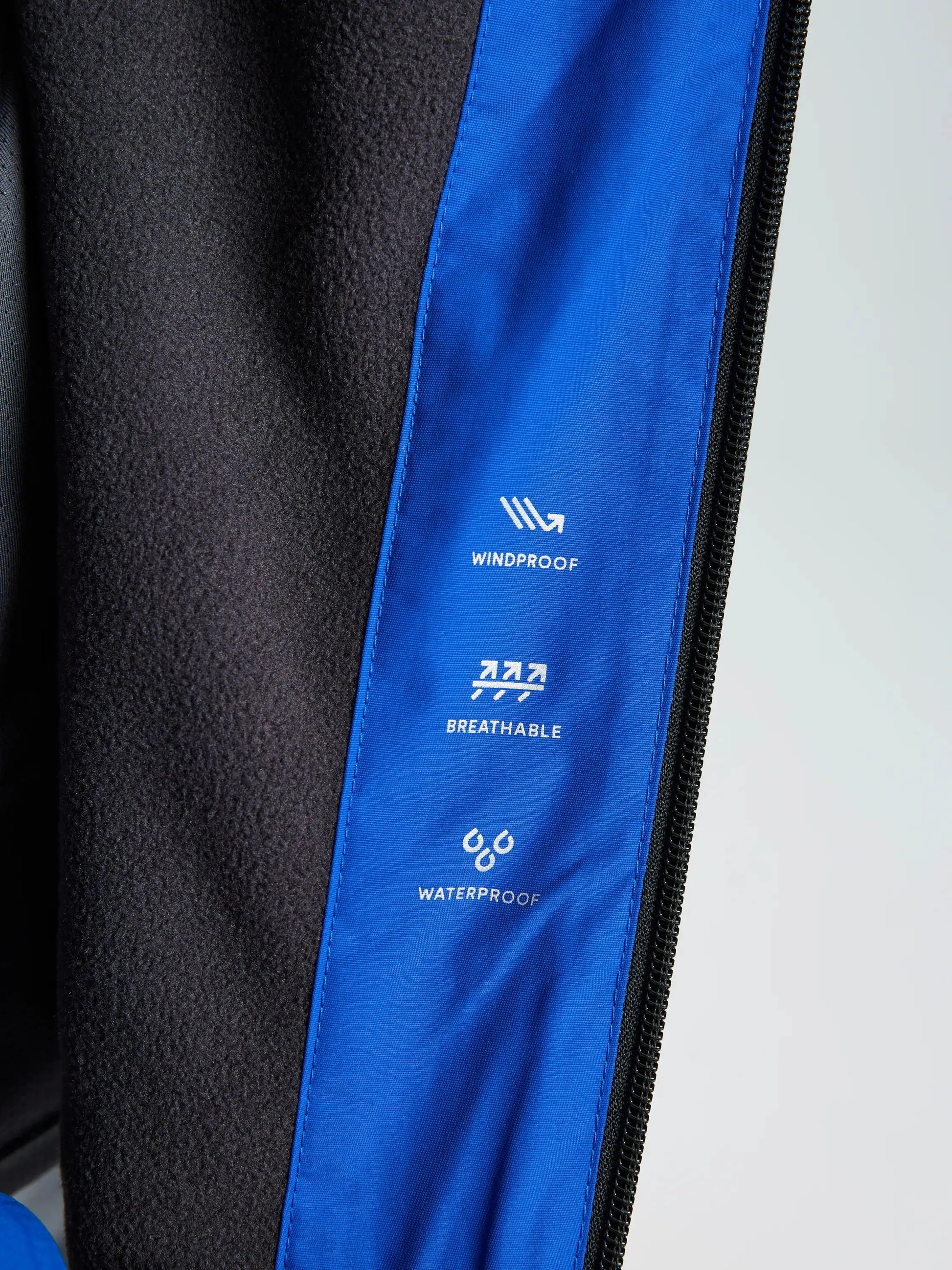 Close-up of the SLAM CREW WARM HOODED JACKET’s inner blue lining, showcasing three icons labeled windproof, breathable, and waterproof to highlight its protective features. The remainder of this black jacket emphasizes its waterproof design.
