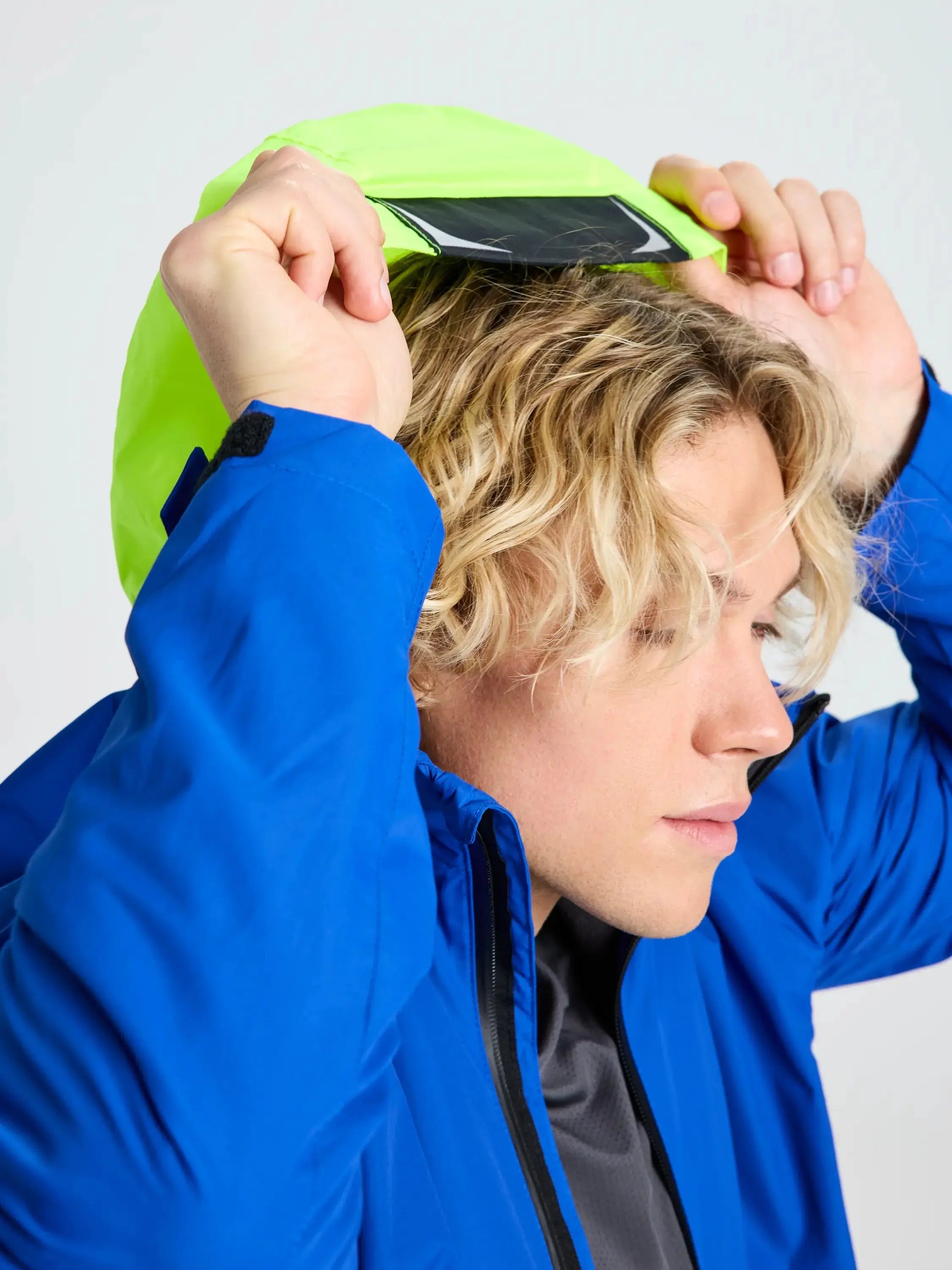A person with wavy blonde hair, wearing the SLAM CREW WARM HOODED JACKET in blue, places a smartphone on their head while lifting the hood, which reveals its lime green polar fleece lining. The background is plain and light-colored.