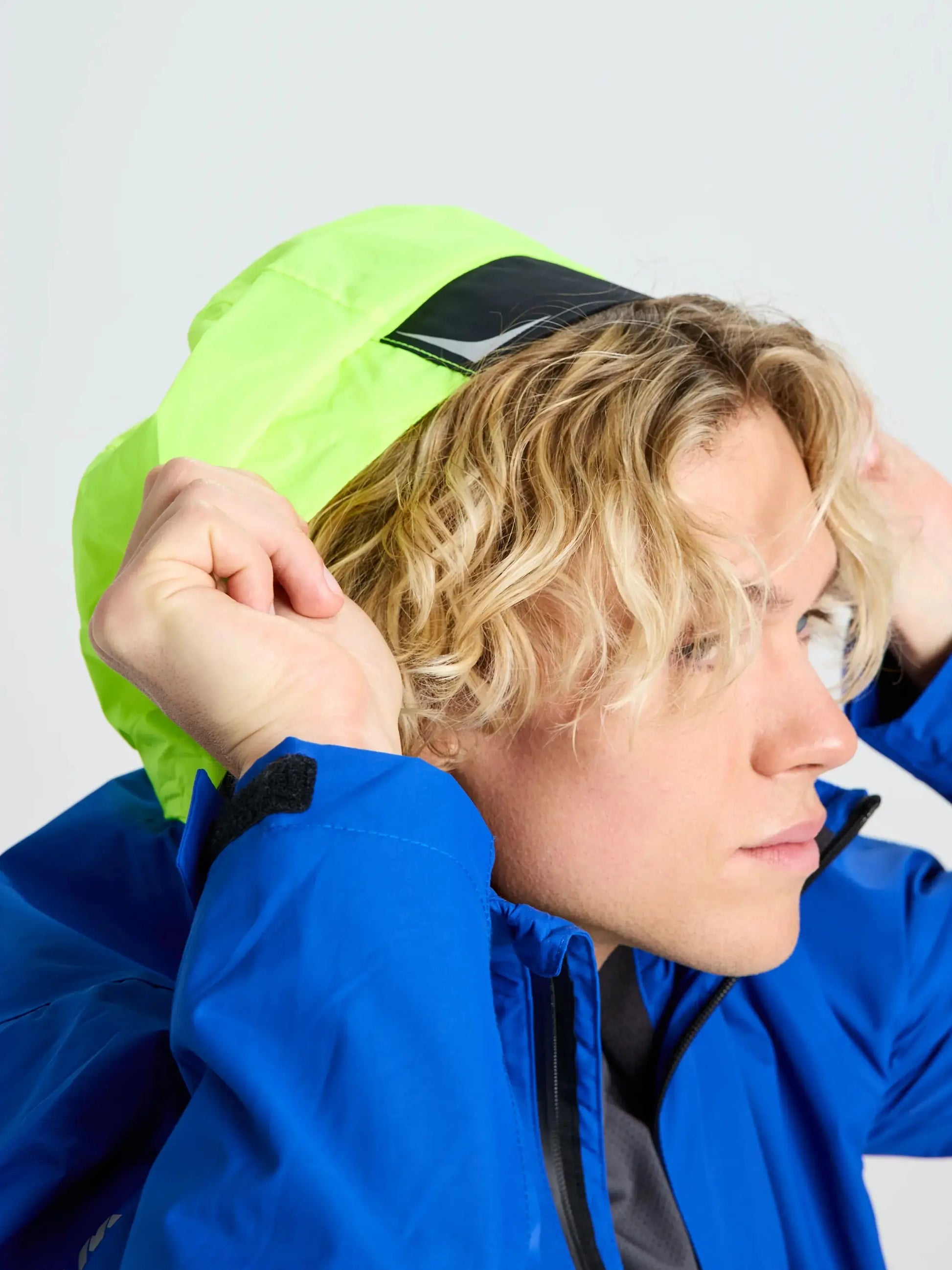 A person with wavy blond hair is shown in side profile against a plain background, wearing the SLAM CREW WARM HOODED JACKET—a bright blue winter sailing jacket—while pulling up its neon yellow hood.