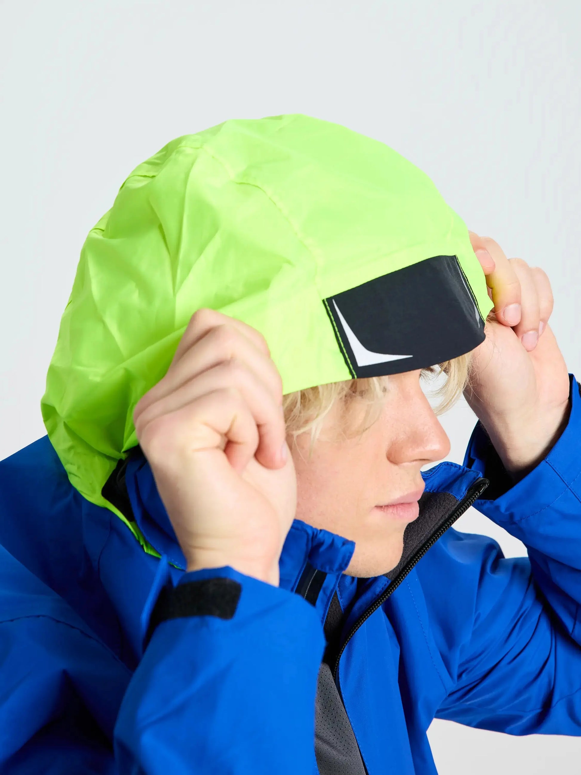 Wearing the SLAM CREW WARM HOODED JACKET in blue, a person lifts the neon yellow hood featuring a black and white rectangular patch on the front; their face is partially visible in profile.