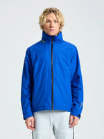 A person with short blond hair wears the SLAM CREW WARM HOODED JACKET in bright blue, paired with light pants, standing against a plain light background and facing the camera with a neutral expression.