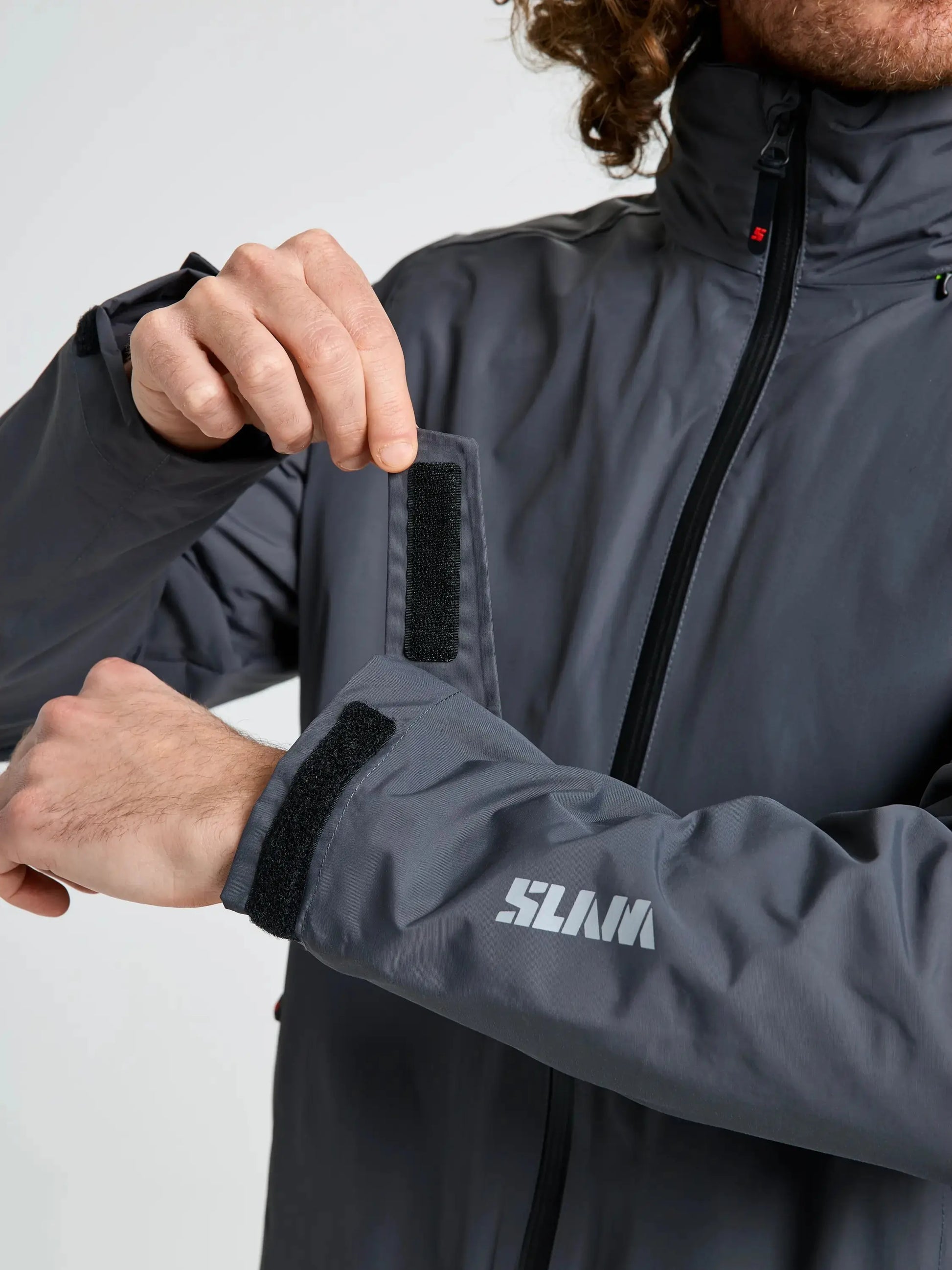 A person adjusts the Velcro strap on the cuff of a gray CREW WARM HOODED JACKET by SLAM, with “SLAM” printed in white on the sleeve. Only the torso, arms, and part of the head are visible, drawing attention to the practical design of this winter sailing jacket.