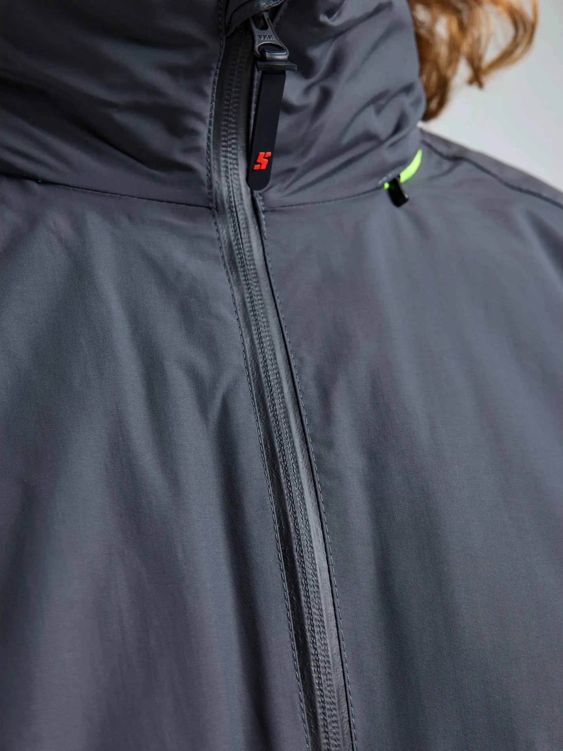 A close-up view of the SLAM CREW WARM HOODED JACKET in gray, showcasing a central zipper, a small red SLAM logo on the zipper pull, and a touch of neon green detail near the collar.