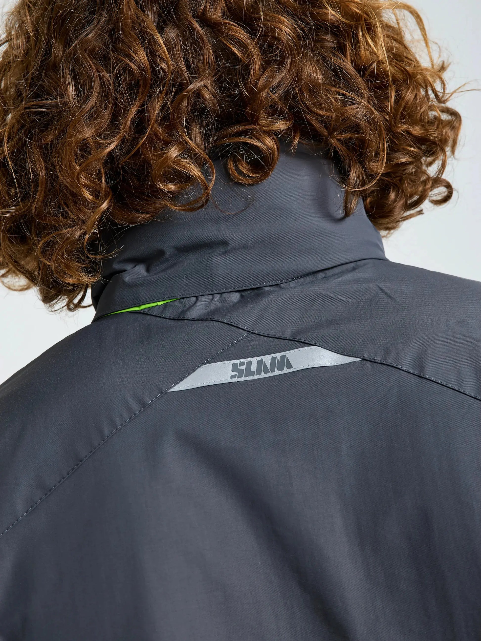The image shows the back of a person with curly brown hair wearing the SLAM CREW WARM HOODED JACKET in dark gray. A reflective SLAM logo is printed near the top center of the jacket’s back.