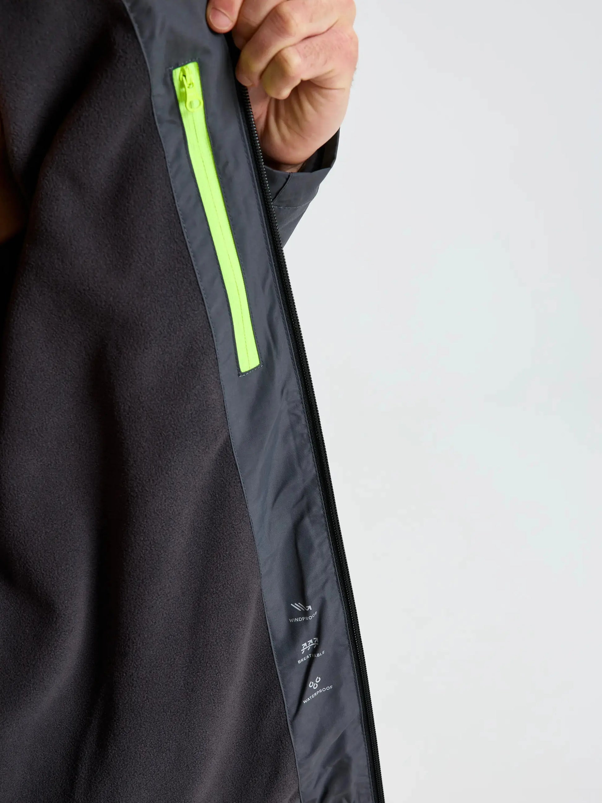 A close-up of the interior of the SLAM CREW WARM HOODED JACKET reveals a neon green zippered pocket on the left side against a gray lining. A hand holds the jacket open, displaying small white care symbols printed near the pocket—features characteristic of premium winter sailing jackets.