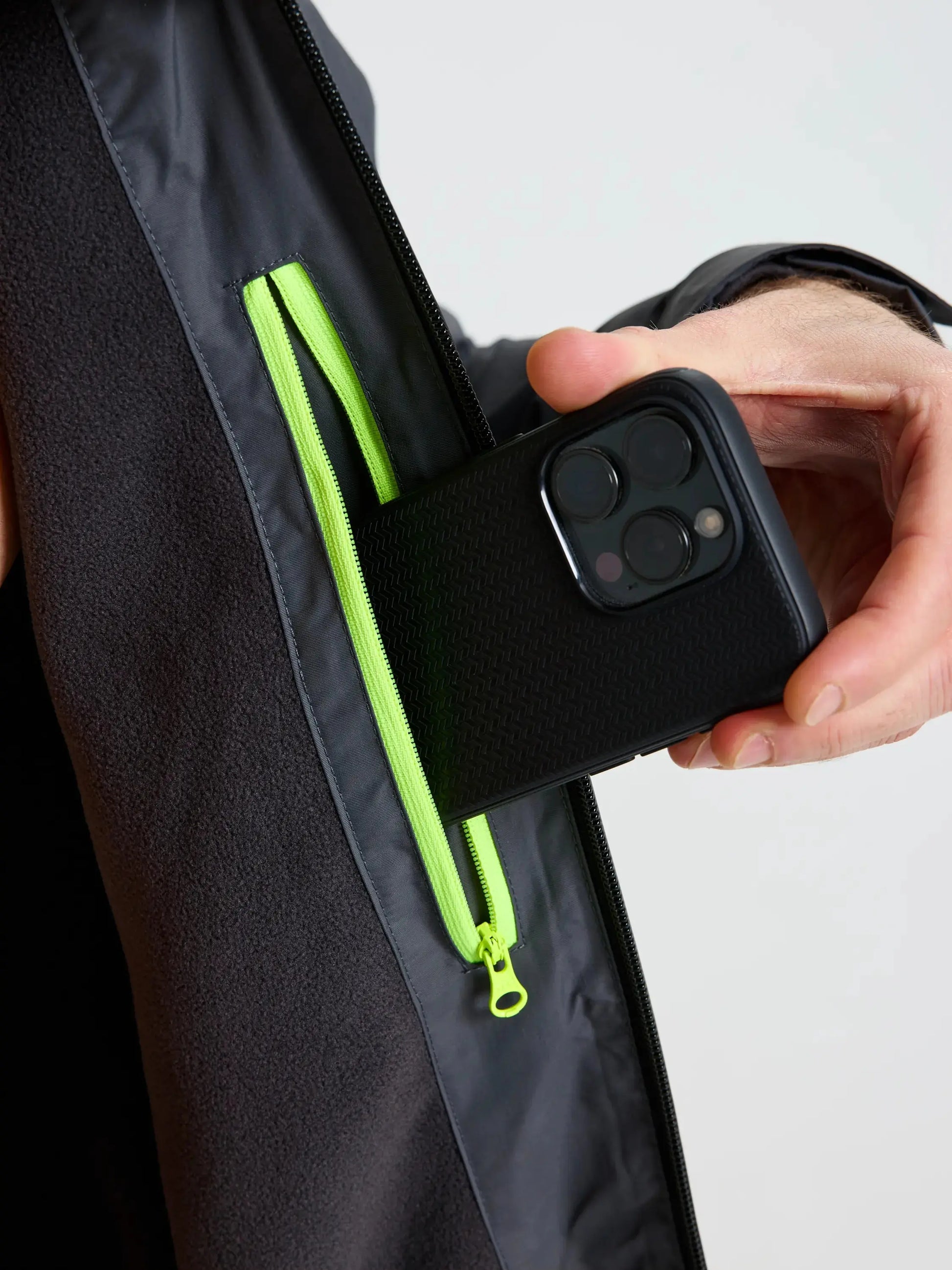 A person places a smartphone with a black textured case into the zippered chest pocket of the SLAM CREW WARM HOODED JACKET, a dark winter sailing jacket featuring a neon green zipper. The background is plain and light-colored.