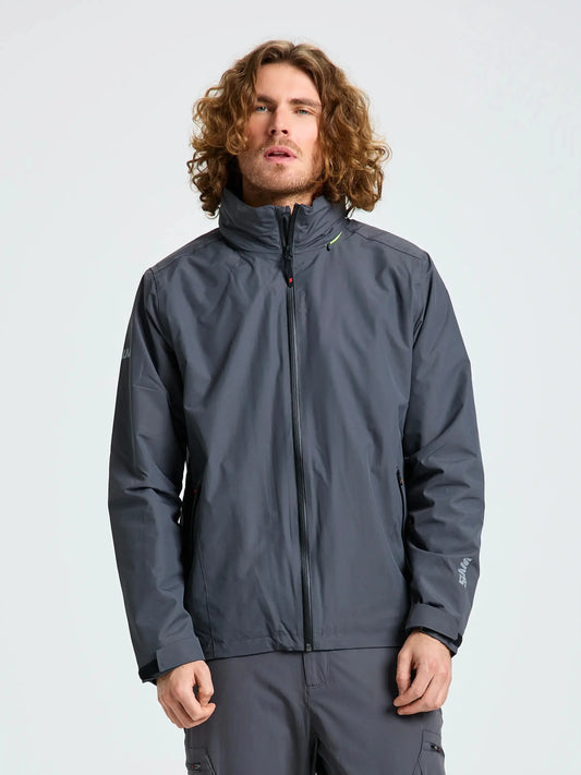 A person with curly hair is standing against a plain background, wearing the SLAM CREW WARM HOODED JACKET—a gray, waterproof jacket inspired by winter sailing jackets. The jacket features a high collar lined with polar fleece, and the person’s hands are relaxed by their sides.
