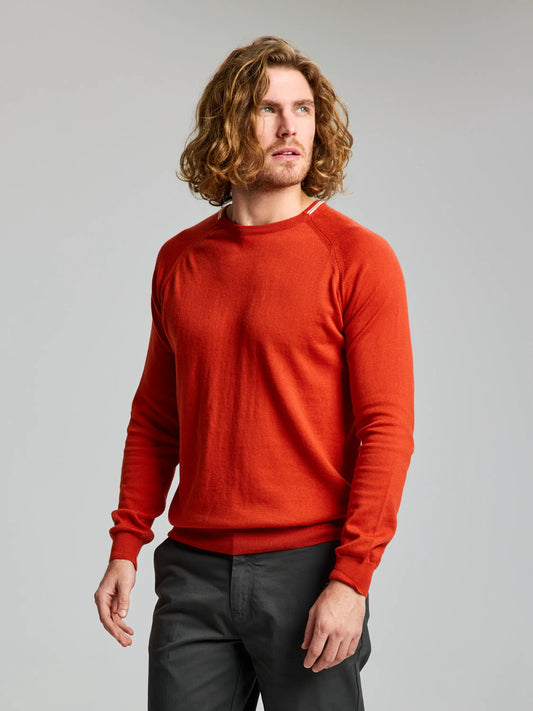 A person with shoulder-length wavy hair is wearing an Ayers Rock JUMPER BOANN CL by SLAM and dark pants. They are standing against a plain light gray background and looking to the side.