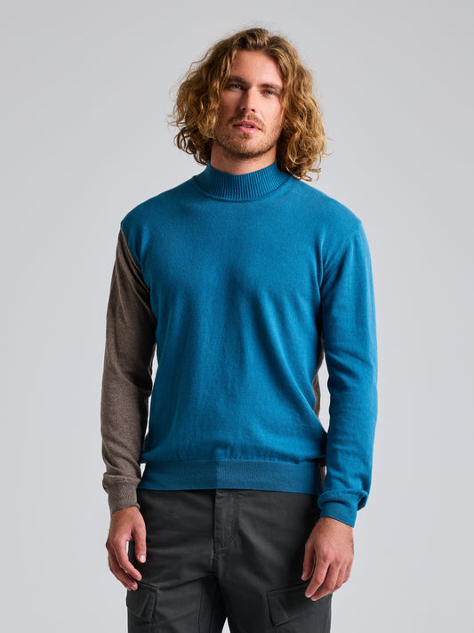 A person with long curly hair is wearing the JUMPER ERSE CL by SLAM, featuring a two-tone design in blue with one brown sleeve, paired with dark green cargo pants and standing against a plain gray background.