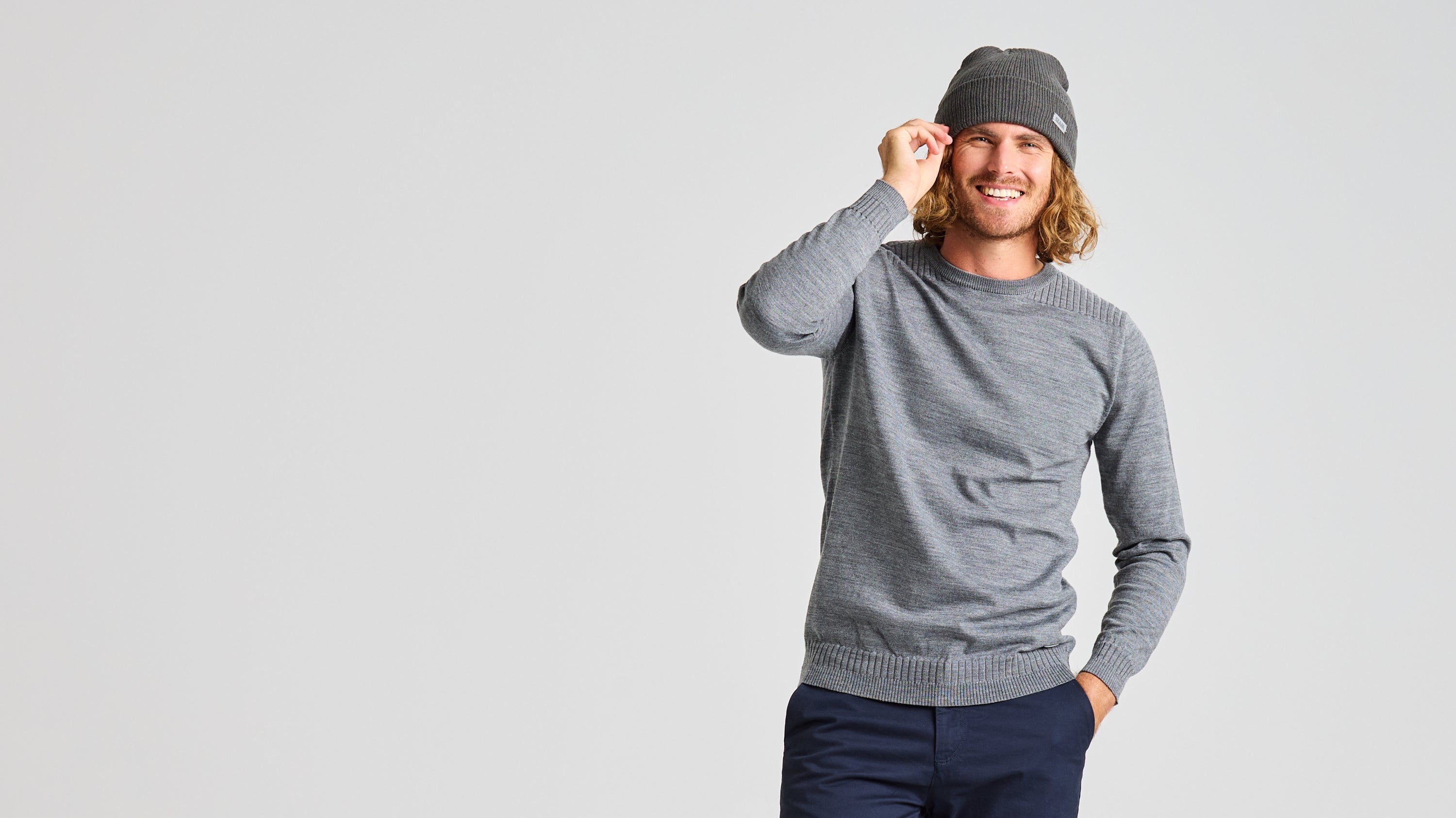 A person with long hair and a beard smiles while wearing a gray SLAM SEAWOOL CREWNECK and a matching beanie, crafted from Merino-blend yarn. Their right hand adjusts the beanie, and their left hand rests in the pocket of their regular-fit dark pants. The background is a plain light gray.