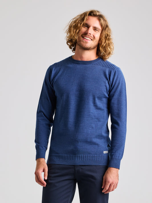 A person with curly hair is wearing a SLAM SEAWOOL CREWNECK sweater in Storm Blue Melange and dark pants, standing against a plain background. This regular fit sweater showcases ribbing details on the collar, cuffs, and hem. The person is smiling with closed eyes.