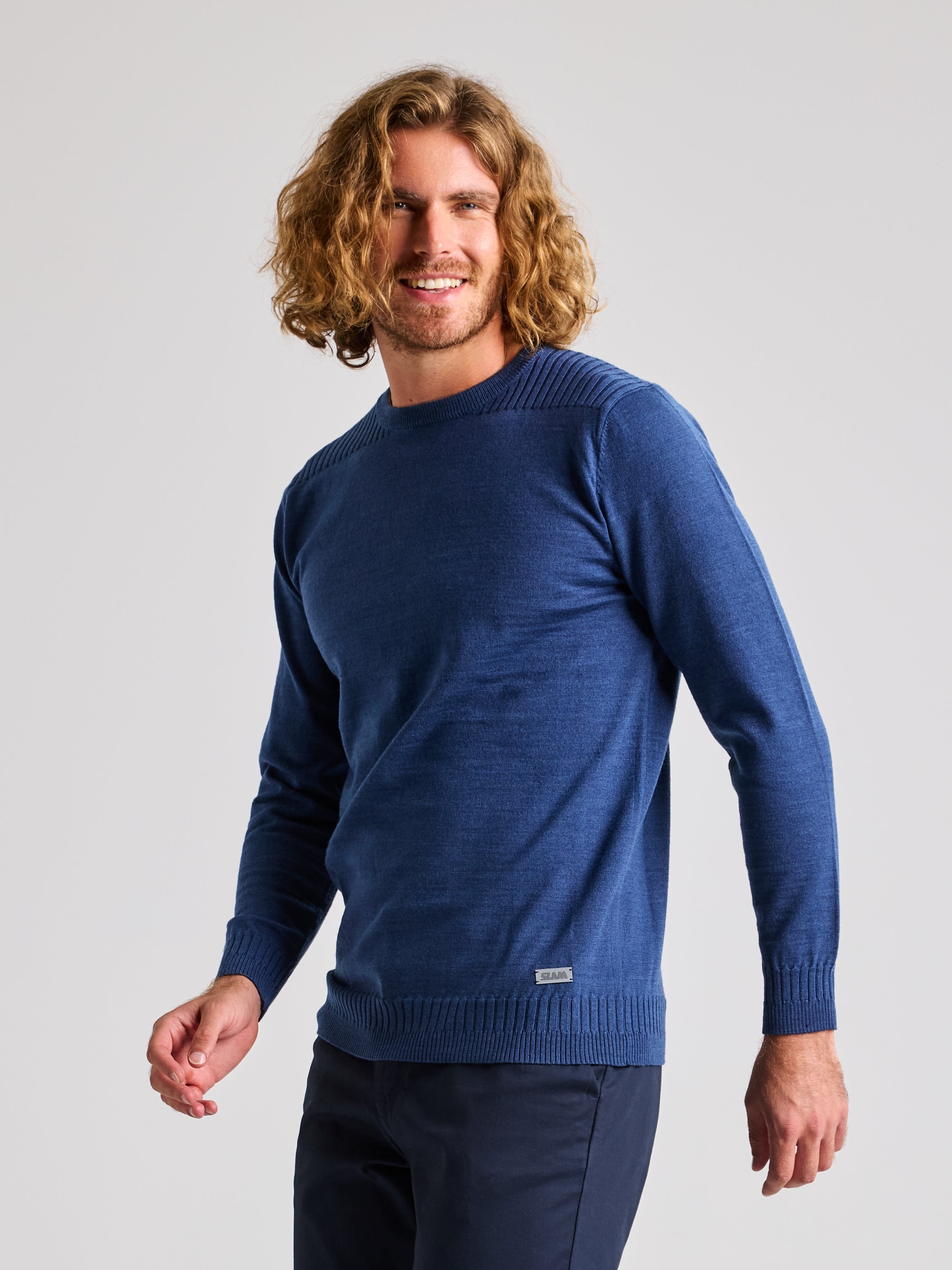 A man with shoulder-length curly hair is wearing a long-sleeved SEAWOOL CREWNECK sweater by SLAM, crafted from Merino-blend yarn in blue, paired with dark, regular fit pants. He stands against a plain gray background, smiling slightly as he looks towards the camera.