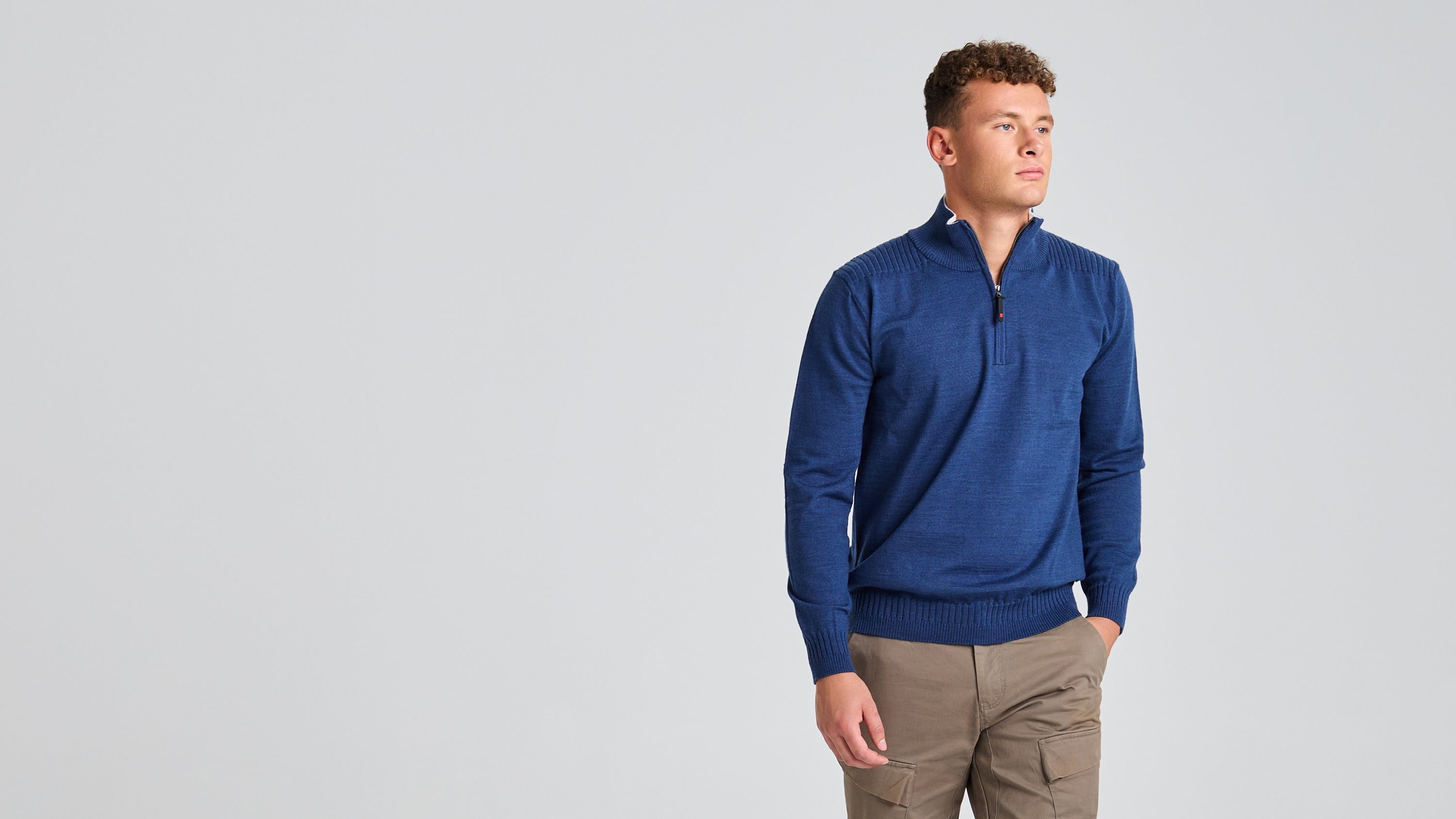 A person wearing a SLAM's SEAWOOL HALF-ZIP in blue and brown pants stands against a plain gray backdrop. With one hand in their pocket, they gaze off to the side, highlighting the sustainable fashion of recycled Seawool Merino-blend yarn.