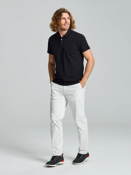 A person with long curly hair is wearing a black polo shirt, SLAM's DECK LIGHT CHINO PANT in light gray stretch cotton twill, and black sneakers featuring orange highlights. They stand against a gray background with their hands in their pockets, gazing to the side.