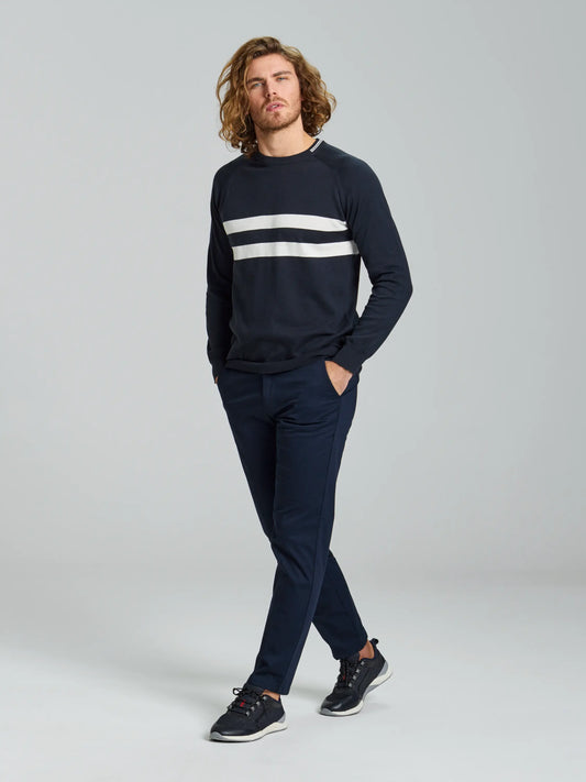 A man with long curly hair strides confidently forward, his hands nestled in his pockets. He wears a dark sweater featuring two white stripes across the chest, paired with SLAM's DECK LIGHT CHINO PANT in stretch cotton twill and black sneakers. The backdrop remains a simple gray.