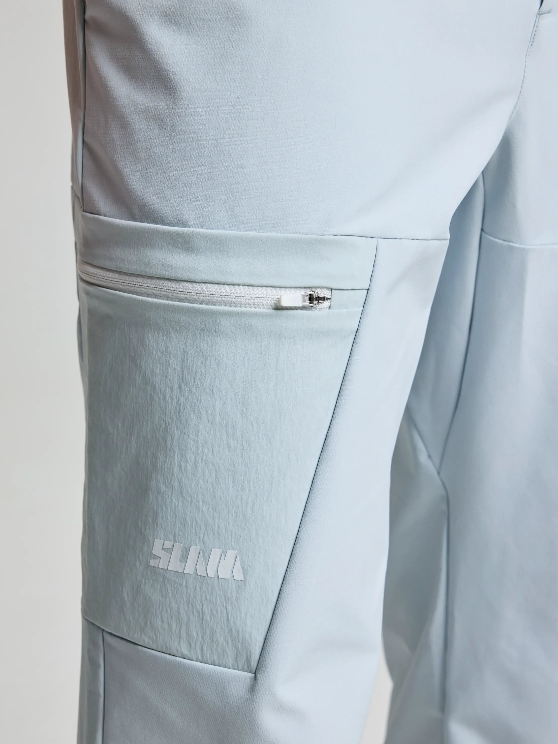 Close-up of SLAM's OD CARGO PANT in light grey, crafted from Double Weave stretch fabric. Feature a textured design with a zippered thigh pocket, white logo below the pocket, and water-repellent properties for versatility.