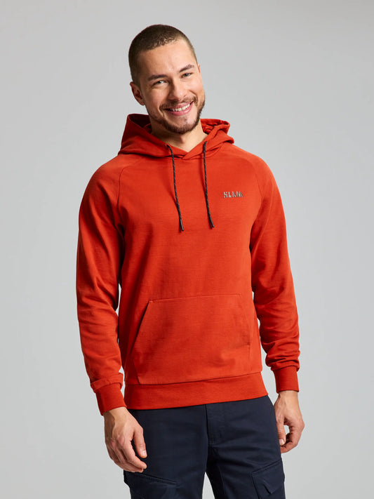 A person with short hair is wearing the DECK HOODIE from SLAM in Ayers Rock color, which features an adjustable hood and a small logo on the chest. The hoodie includes a front pouch pocket, and paired with dark pants, they smile while standing against a plain background.