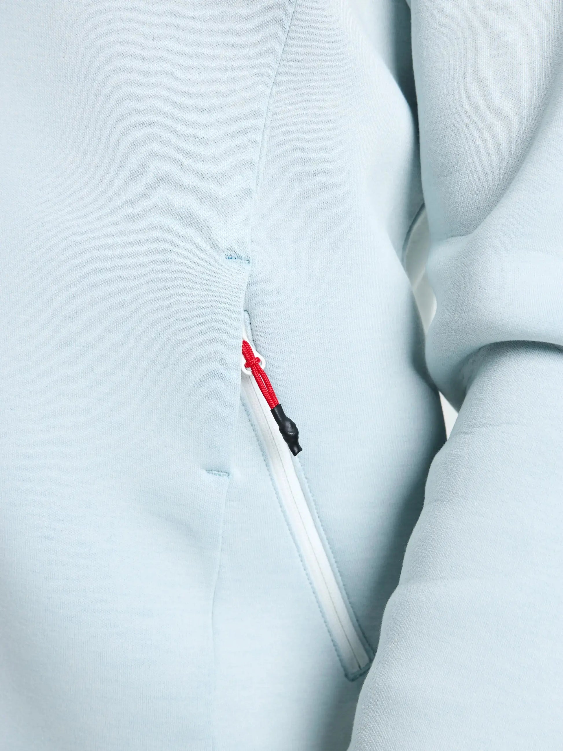 Close-up of the SLAM ACT FULL-ZIP HOODIE in light blue, crafted from stretchy scuba fabric. The hoodie features a white zippered pocket with a zipper pull that is white and detailed with a red string and black tip. A portion of the sleeve is visible on the right side of the image.