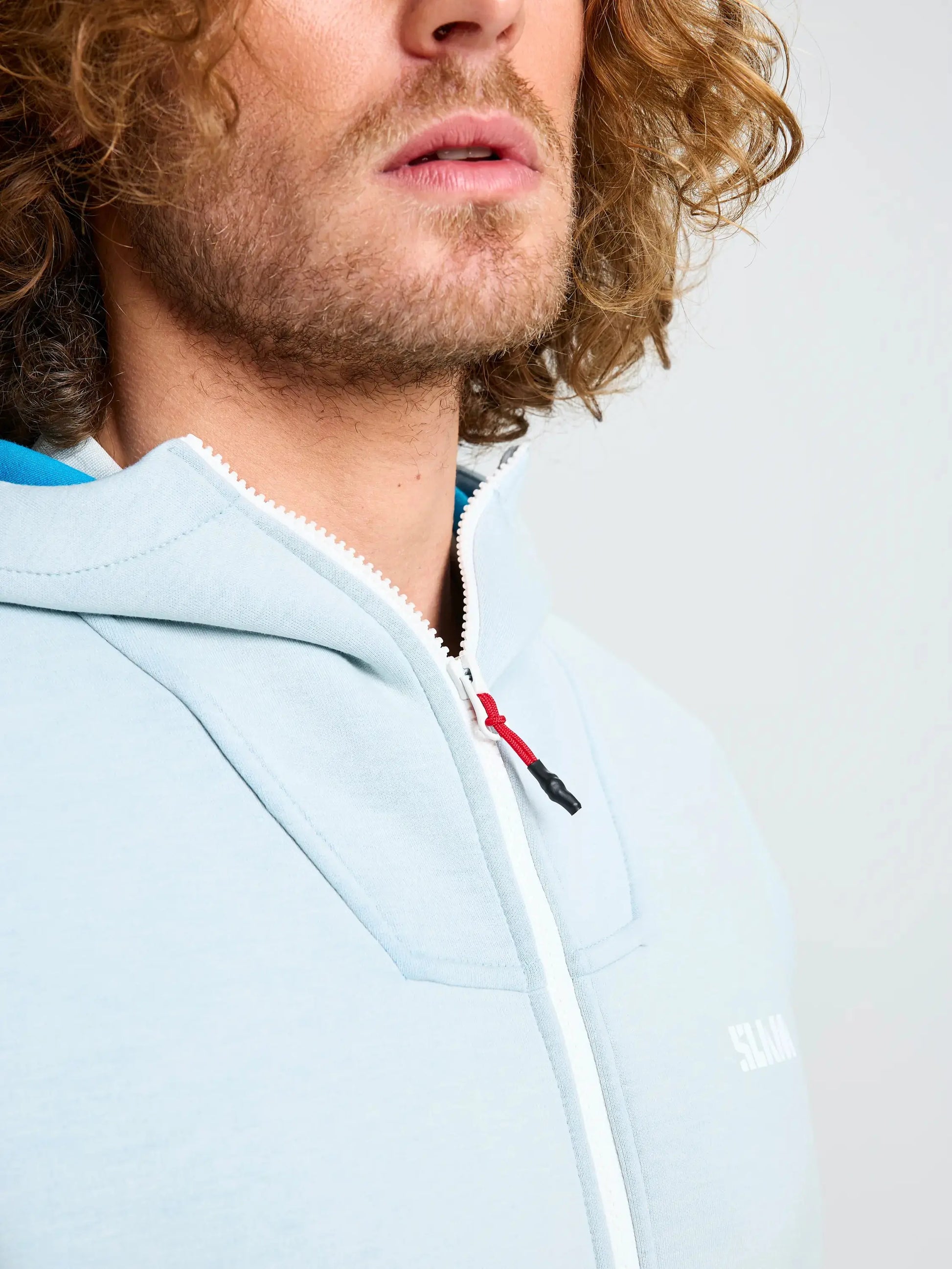 A person with curly hair is wearing the SLAM ACT FULL-ZIP HOODIE in light blue, crafted from stretchy scuba material. The hoodie features a white zipper with a small red accent on the zipper pull and offers a regular fit, standing out against the simple light gray background.