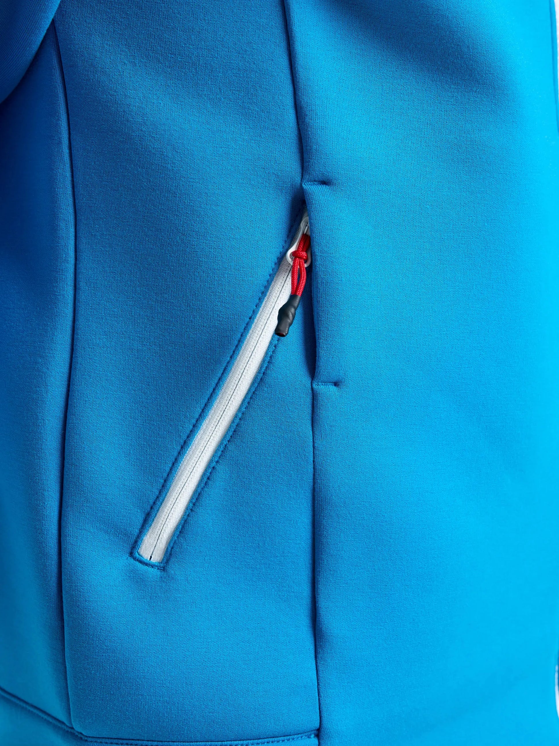 Close-up of the SLAM ACT FULL-ZIP HOODIE in blue, showcasing a side pocket with a gray zipper and a small red zipper pull. Made from stretchy scuba material for enhanced comfort, the image highlights the smooth texture, precise seam details, and regular fit of this stylish hoodie.