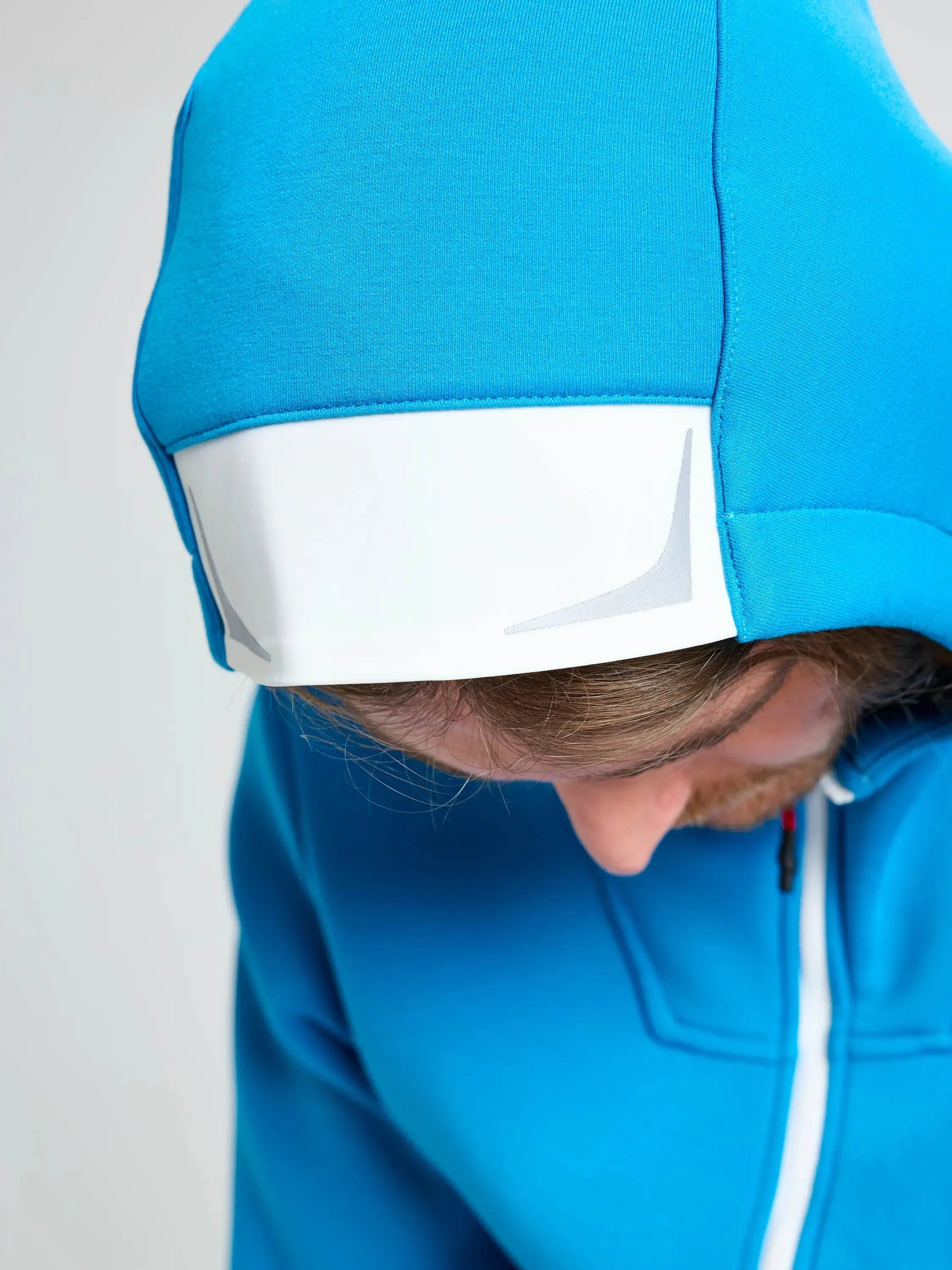 A person wears the SLAM ACT FULL-ZIP HOODIE in bright blue, made from stretchy scuba material, featuring a white rectangular patch on the hood. They are looking downward, mostly hiding their face, against a plain light-colored background.