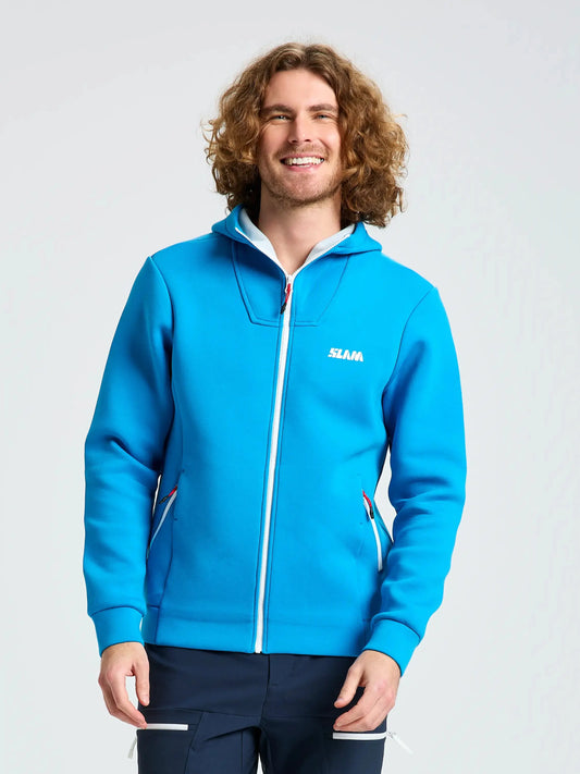 A person with curly hair is wearing a SLAM ACT FULL-ZIP HOODIE in bright blue, made from stretchy scuba material and featuring a white “SLAM” logo on the chest. The regular fit hoodie includes a hood and zippered pockets. The person is standing against a plain, light background and smiling.