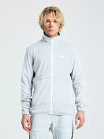 A person with light curly hair wears the SLAM ALTURA FZ LGT GRID FLEECE jacket and matching CLIFF GREY pants, featuring two zippered pockets and a small white chest logo, standing against a plain light background.