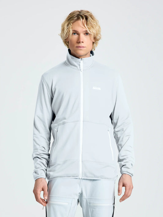 A person with light curly hair wears the SLAM ALTURA FZ LGT GRID FLEECE jacket and matching CLIFF GREY pants, featuring two zippered pockets and a small white chest logo, standing against a plain light background.