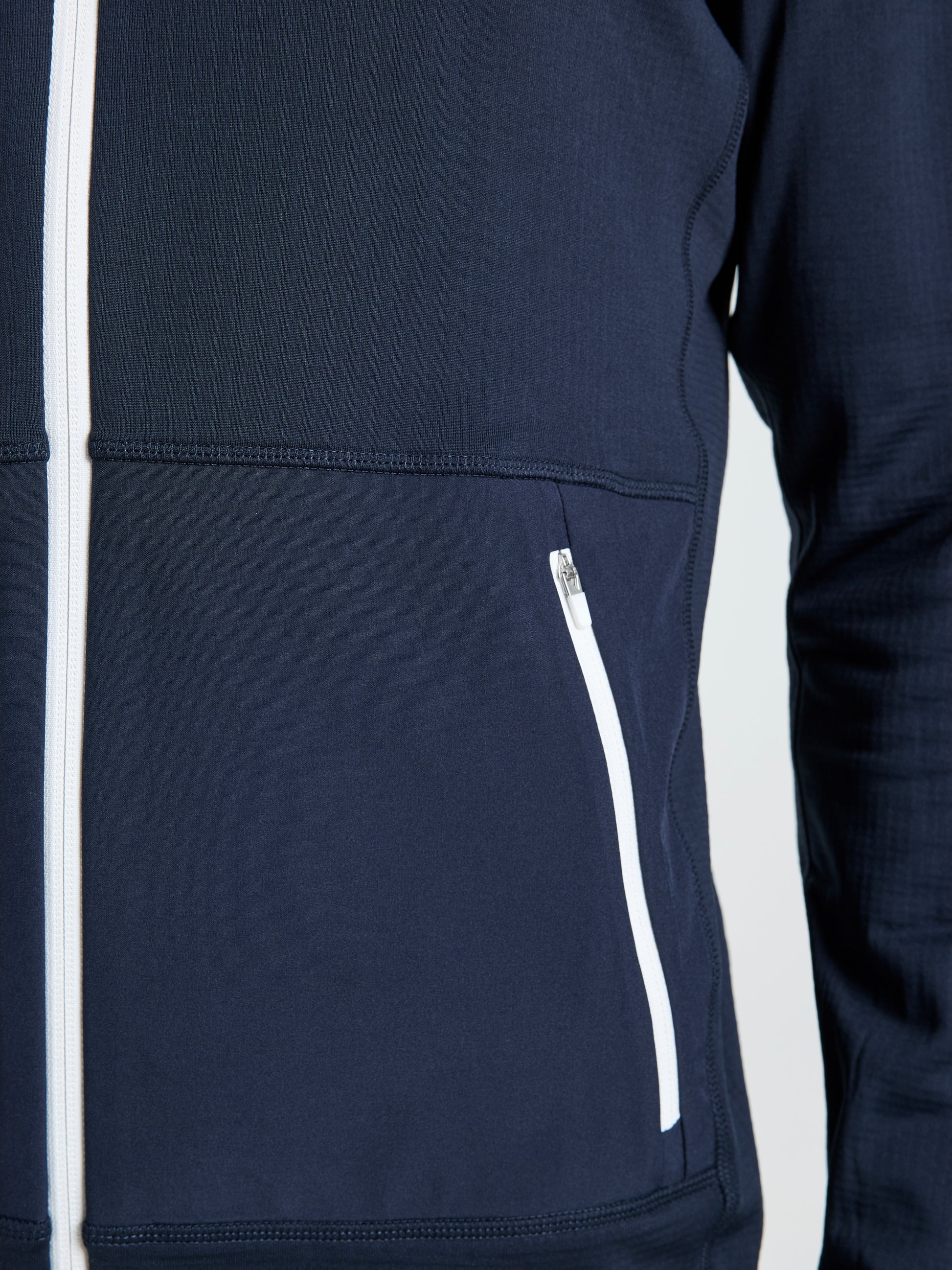 Close-up of a person wearing the SLAM ALTURA FZ LGT GRID FLEECE, a dark blue technical jacket with white front and pocket zippers—perfect for sailing. Only part of the torso and right arm are visible.