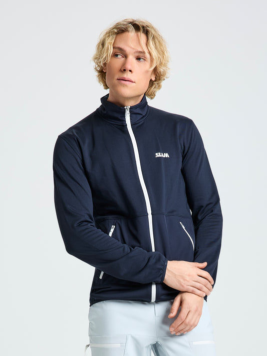 A person with wavy blond hair wears the SLAM ALTURA FZ LGT GRID FLEECE—a navy blue lightweight technical zip-up jacket with white accents and "SLAM" on the chest—standing against a plain light gray background.