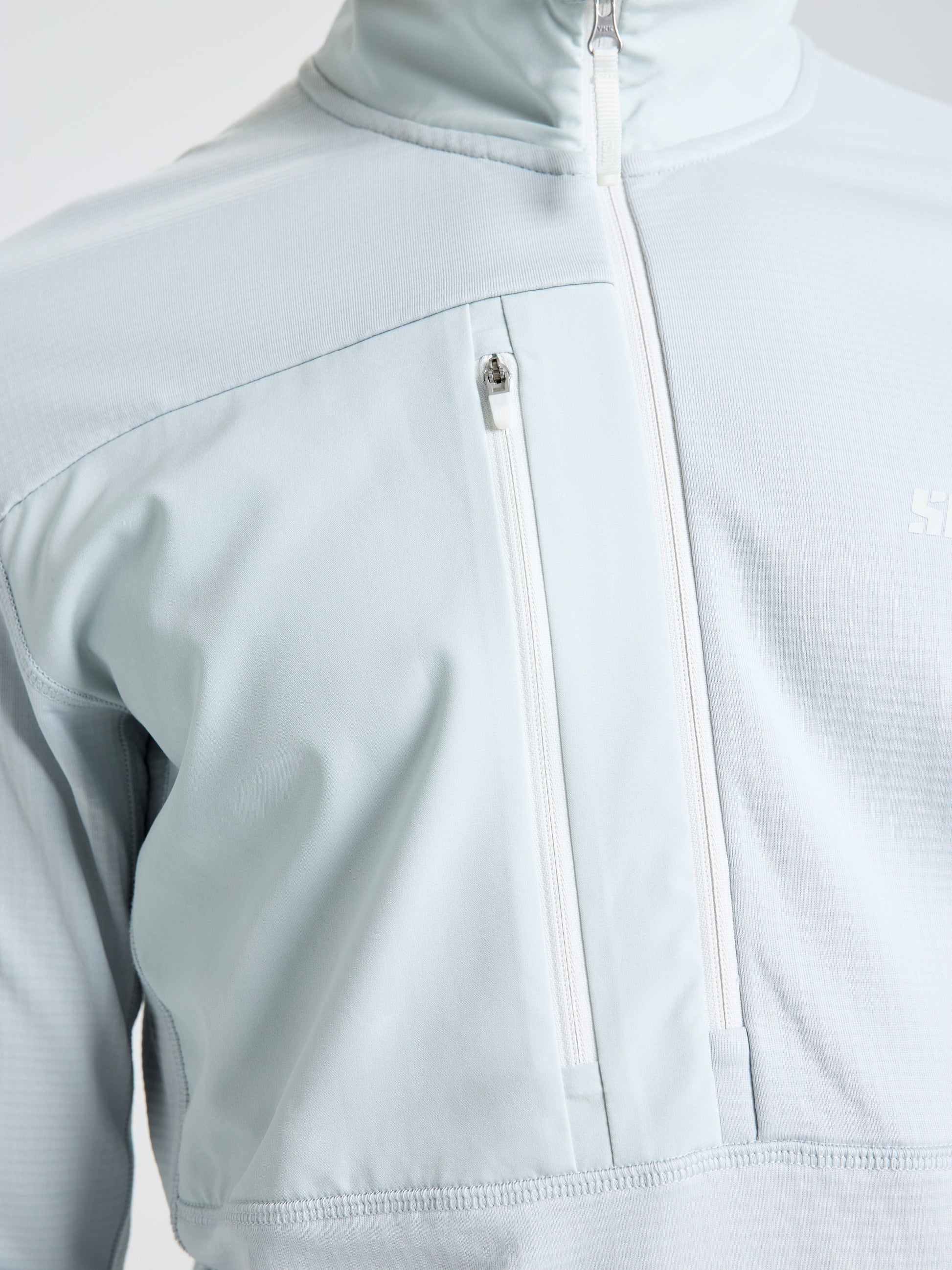 A close-up view of the SLAM ALTURA HZ LGT GRID FLEECE, a light gray zip-up jacket with a high collar, vertical chest pocket with white zipper, and subtle textured pattern for enhanced thermal regulation as a versatile mid layer.