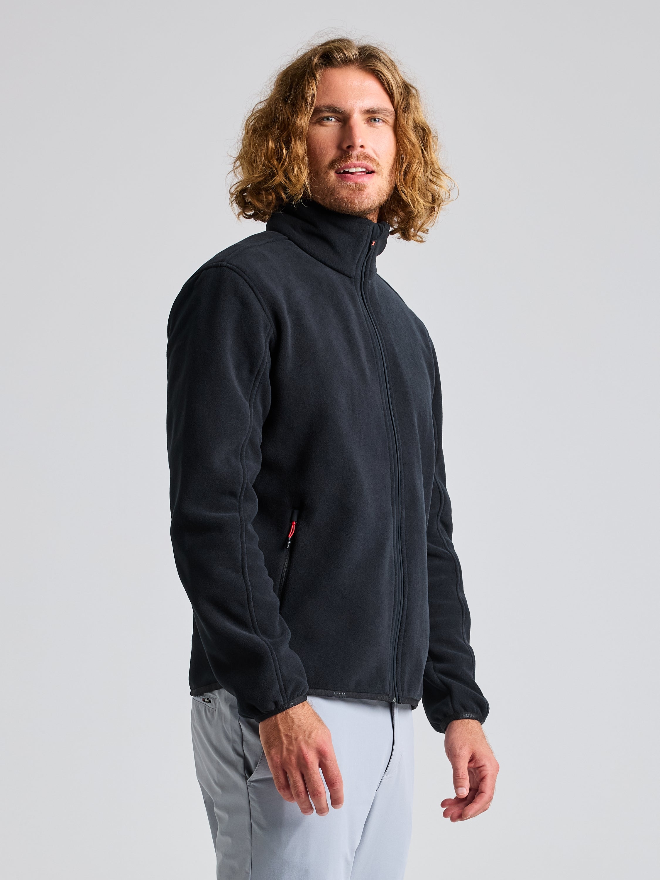 A person with long curly hair is wearing a sleek black DECK FLEECE jacket by SLAM, crafted from recycled microfibre, along with light gray pants. They stand against a plain grey background, facing slightly to the side.